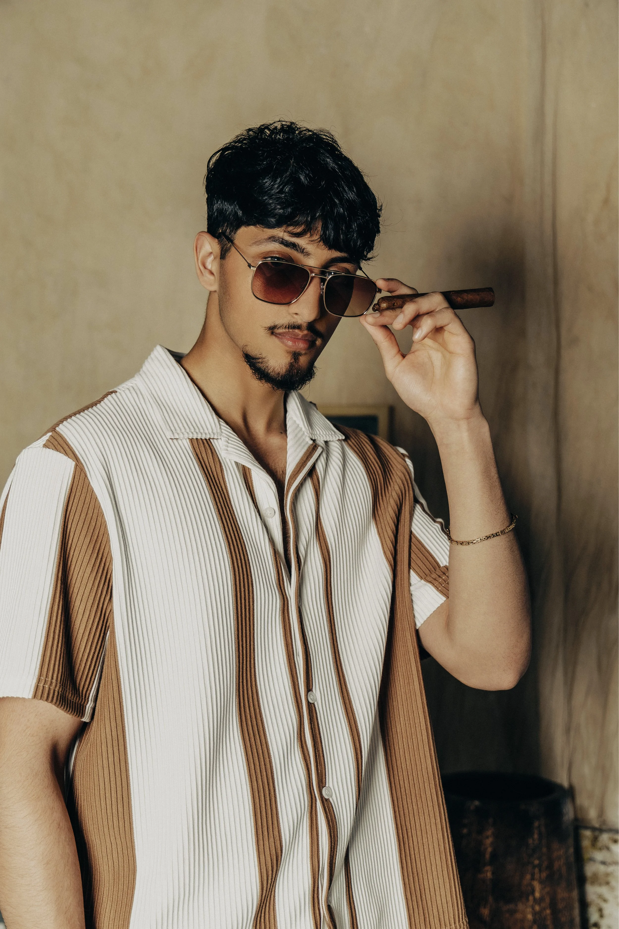 A young man with dark hair, facial hair, wearing sunglasses, a striped shirt, and holding a cigar, standing indoors.