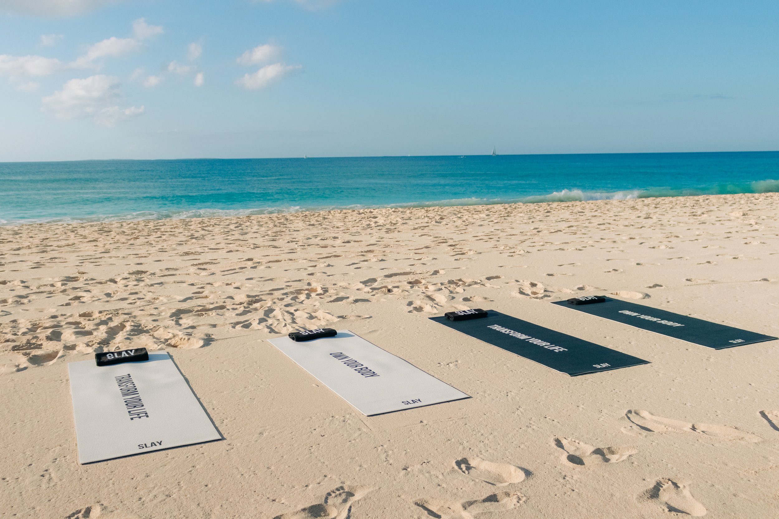 Four yoga mats on sandy beach facing the ocean, with blue sky and a few clouds, and sailboats on the water in the background.