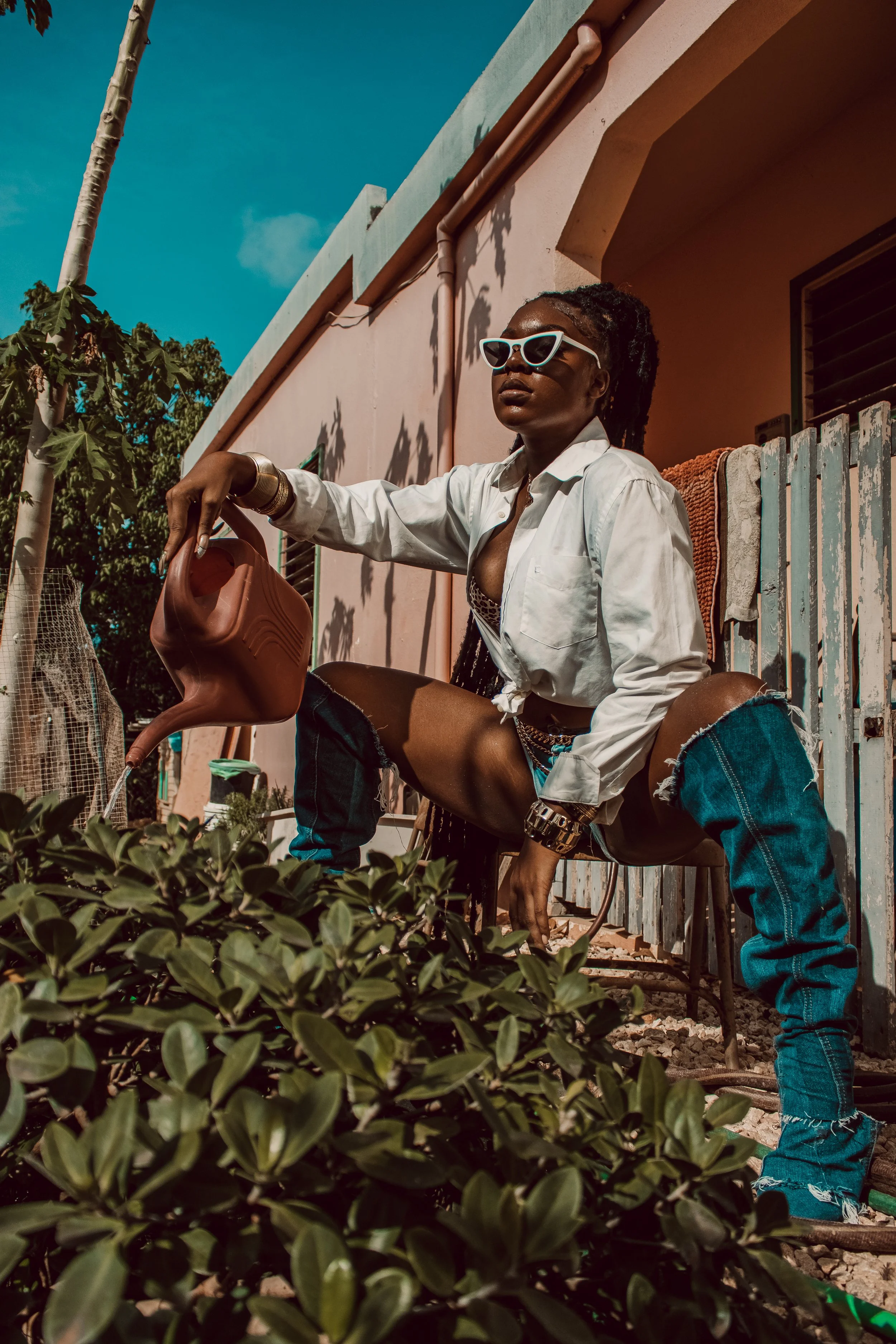 A woman with dark skin wearing sunglasses, a white shirt, and ripped jeans is sitting outdoors, watering plants with a watering can. She has multiple jewelry pieces and has her shirt unbuttoned at the top. The setting includes a pink building, a bamb