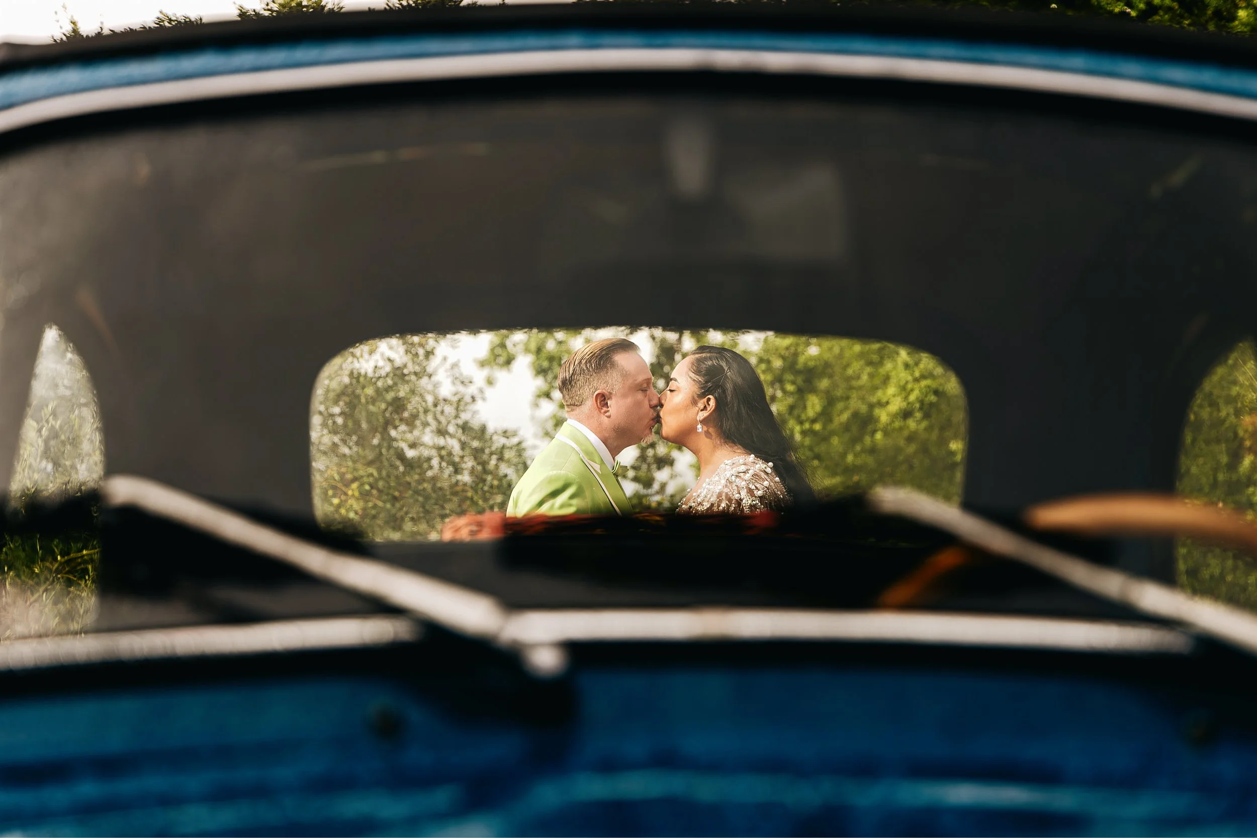 A bride and groom sharing a kiss, viewed through the window of a vintage car, with a background of trees and greenery.