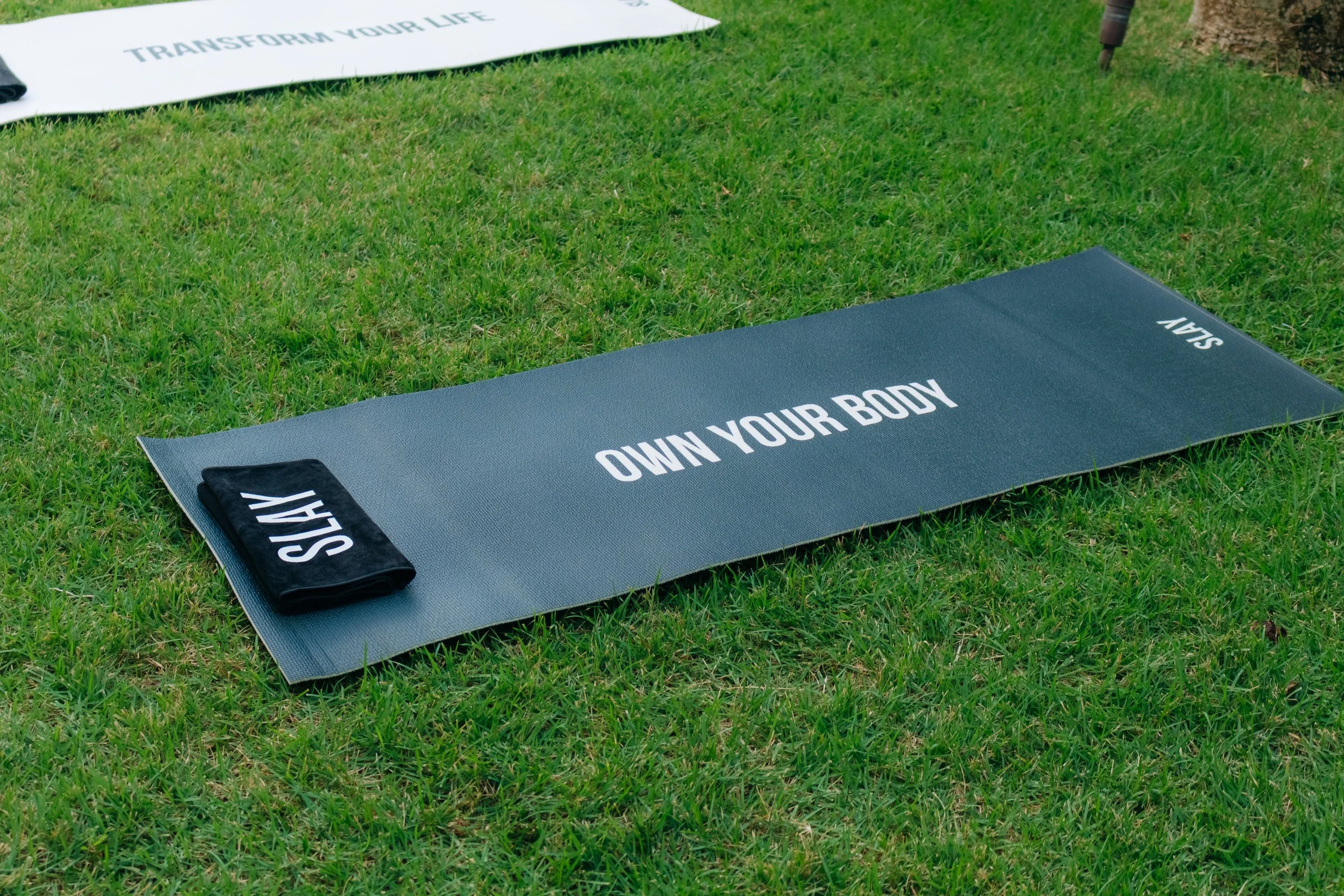A gray exercise mat on green grass with the words 'OWN YOUR BODY' printed in white, and black towels with 'STAY' and 'SAY' written on them.