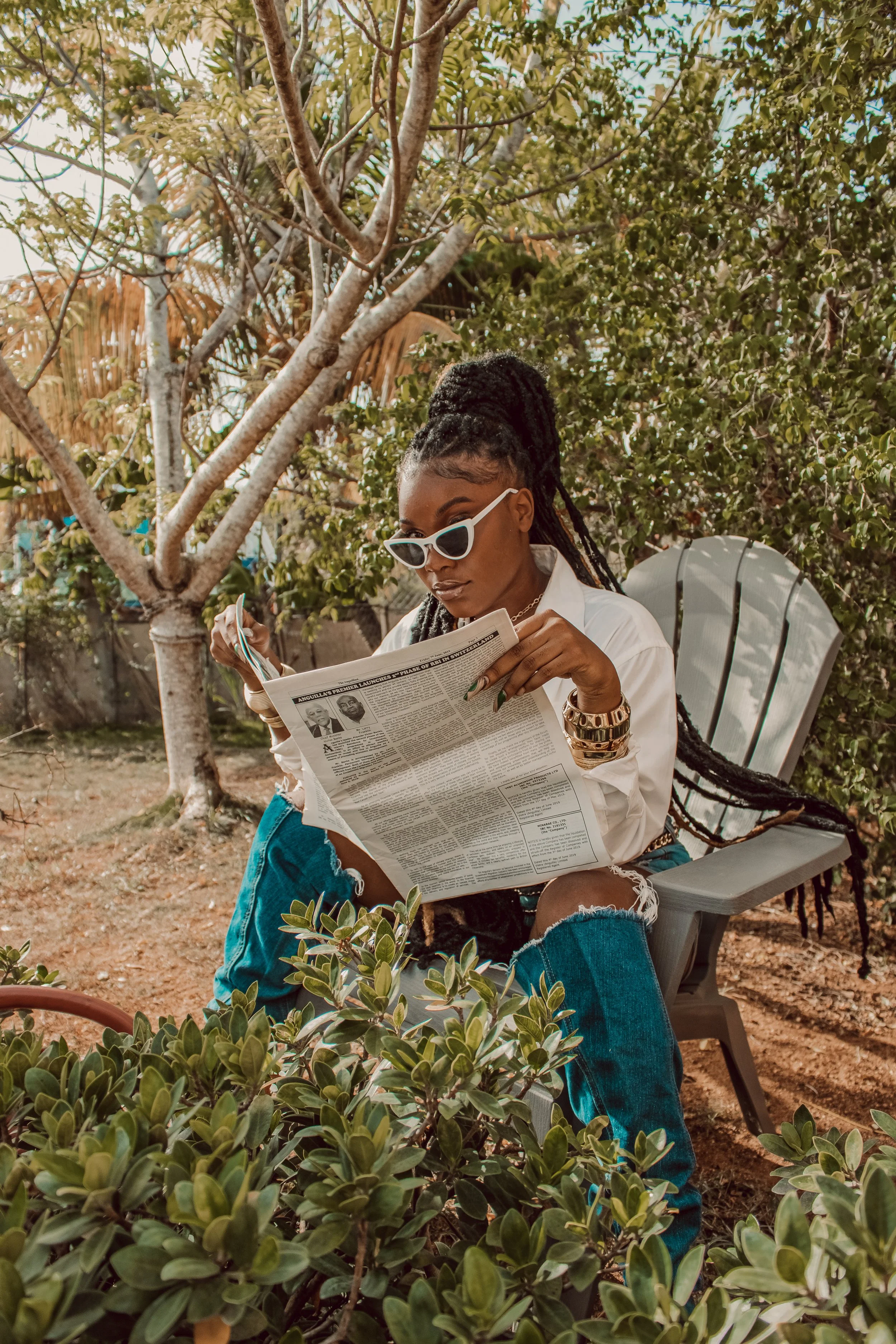 A woman with long braided hair, wearing white sunglasses, a white shirt, and distressed jeans, sits outdoors on a white chair reading a newspaper surrounded by green plants and trees.