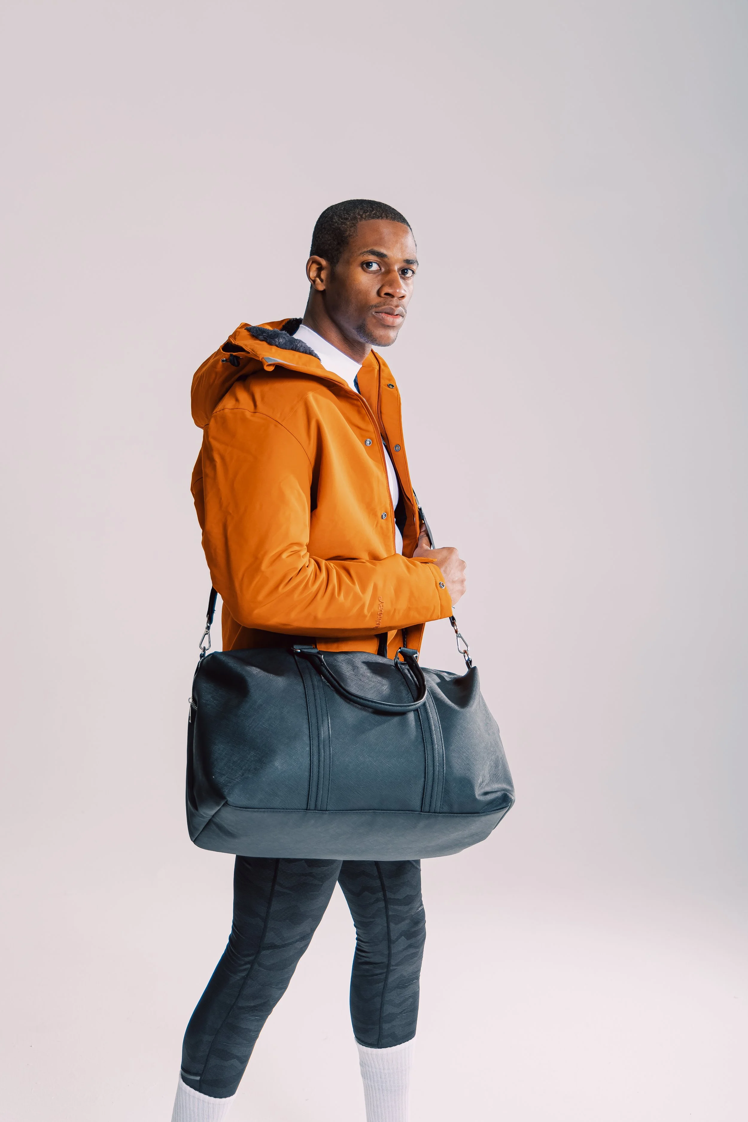 A young man in an orange jacket holding a black duffel bag over his shoulder, standing against a plain light background.