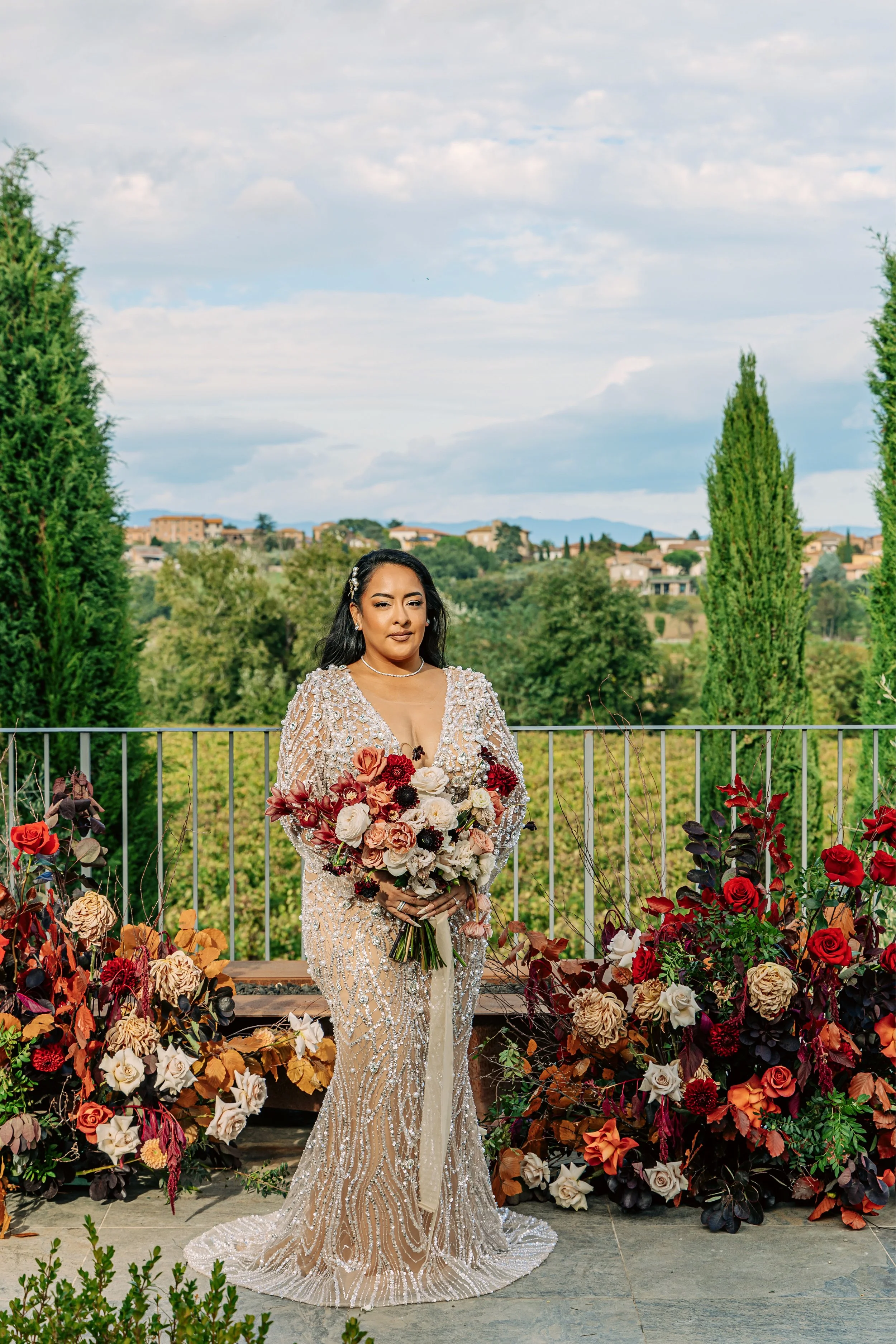 A woman in a sparkling, beaded, cream-colored gown holds a large bouquet of roses, standing outdoors on a balcony decorated with colorful flowers, with a scenic view of trees and houses in the background.