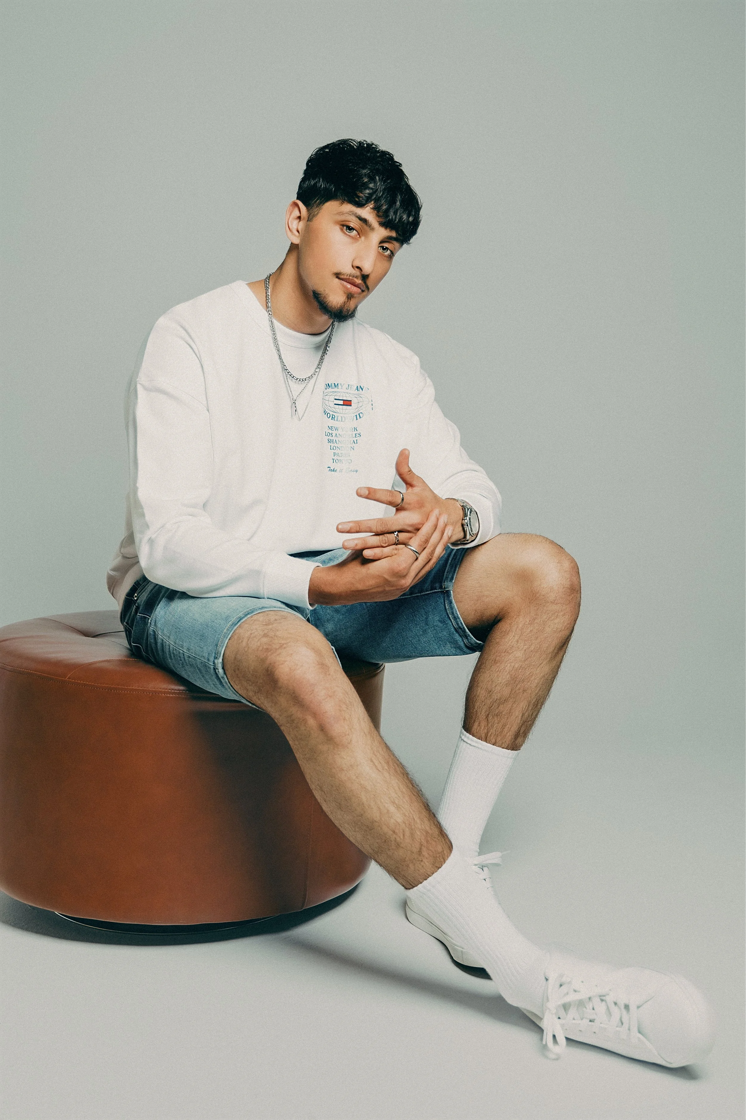 Young man dressed casually in a white long-sleeve shirt, denim shorts, white socks, and sneakers, sitting on a brown leather ottoman against a plain light gray background.
