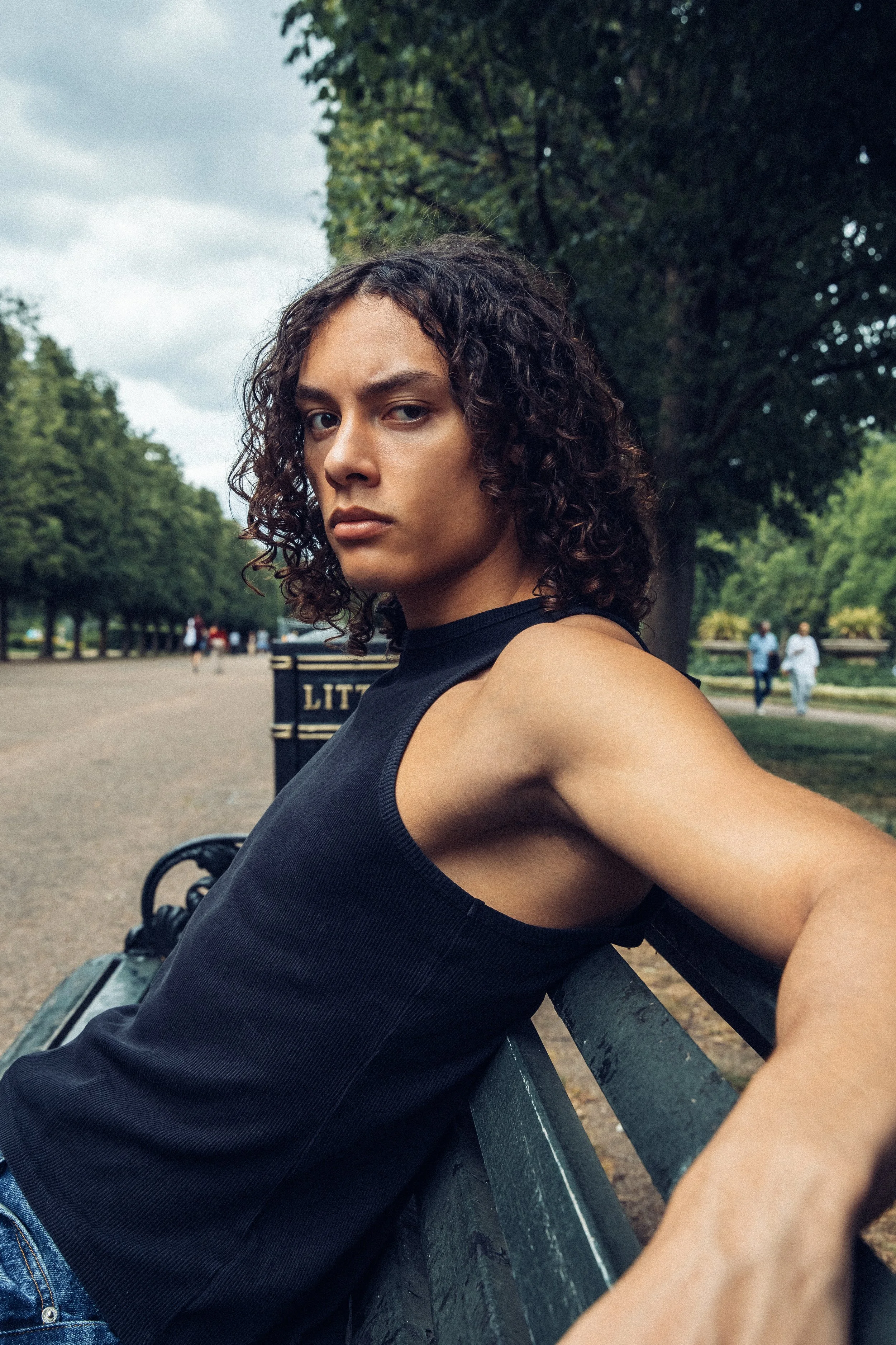 A woman with curly dark hair sitting on a park bench, looking at the camera with a serious expression. She is wearing a black sleeveless top and appears to be in a park with trees and a cloudy sky in the background.