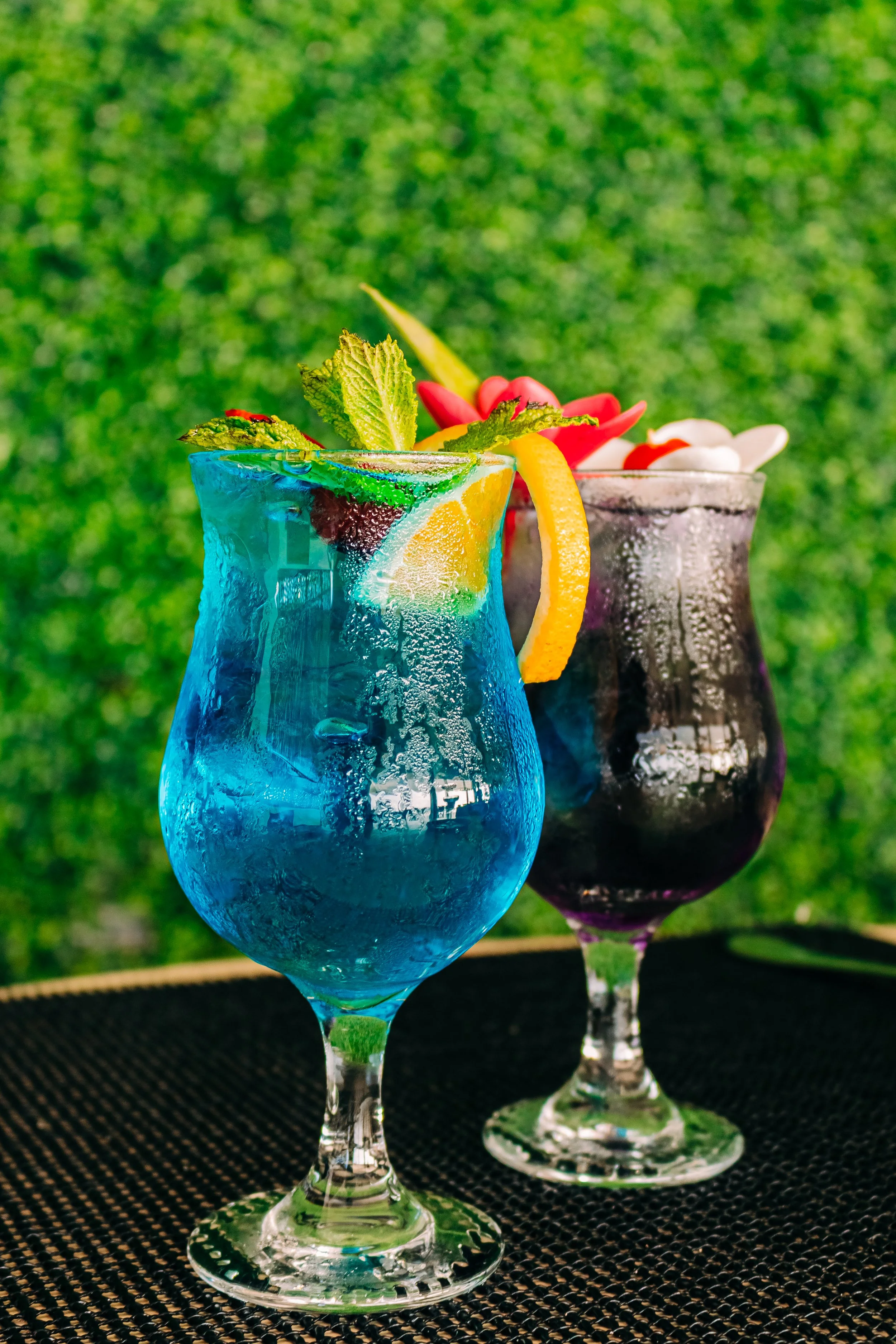 Three colorful cocktails in elegant glasses with fruit garnishes, set outdoors on a black surface with a blurred green background.