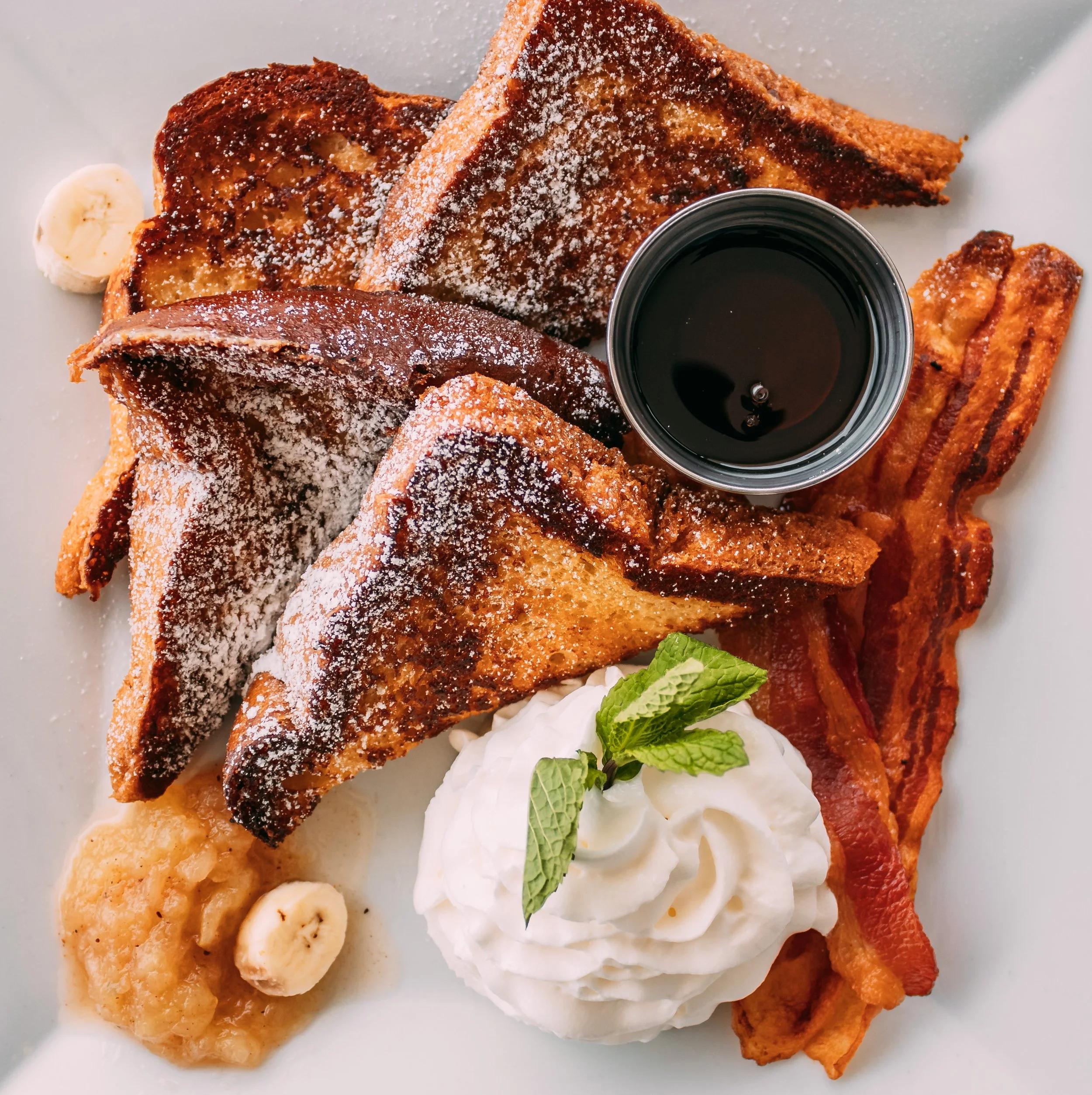 Assorted breakfast items including French toast dusted with powdered sugar, banana slices, a small cup of syrup, whipped cream, and a sprig of mint on a white plate.