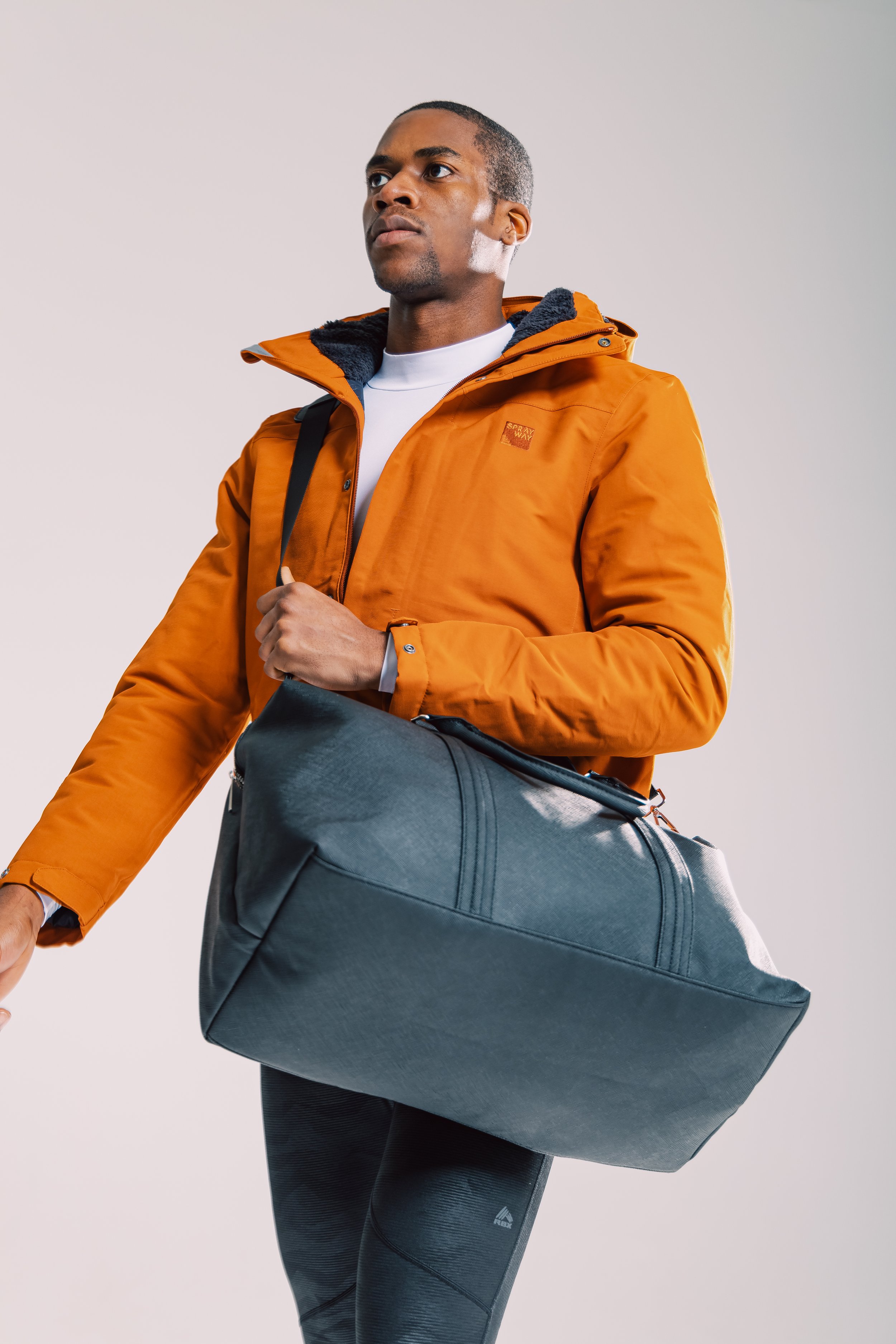 Man wearing an orange jacket and carrying a black duffel bag against a plain background.