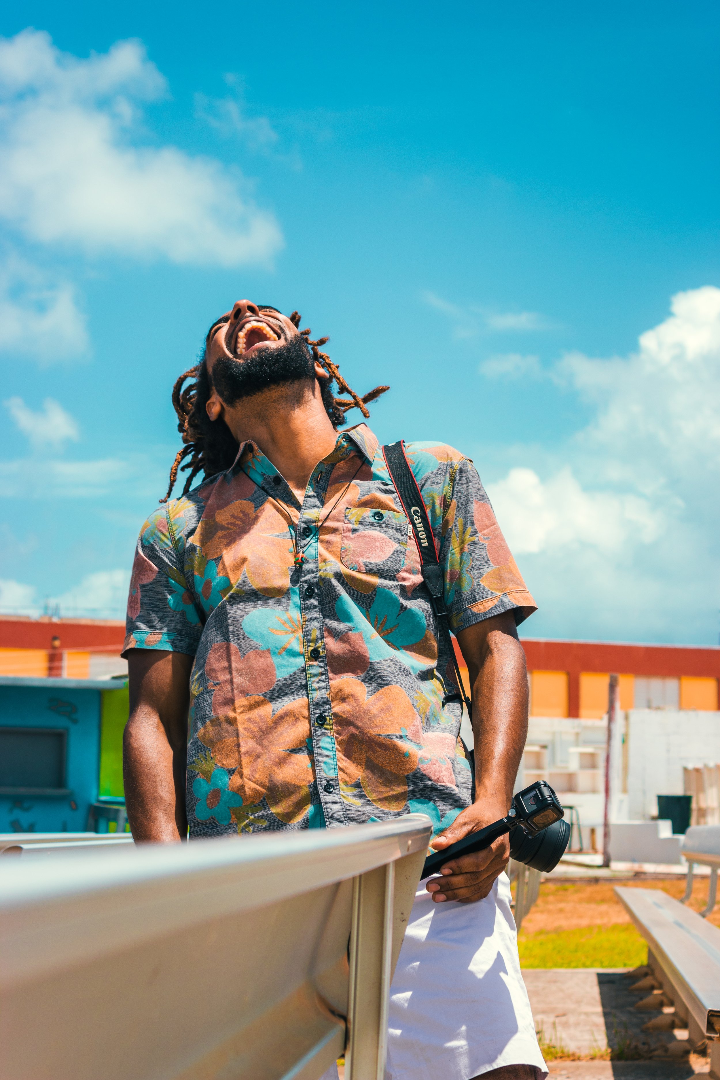 A man with dreadlocks wearing a colorful, floral short-sleeve shirt is outdoors, smiling and laughing, holding a camera and a phone, with a school of colorful painted buildings behind him.