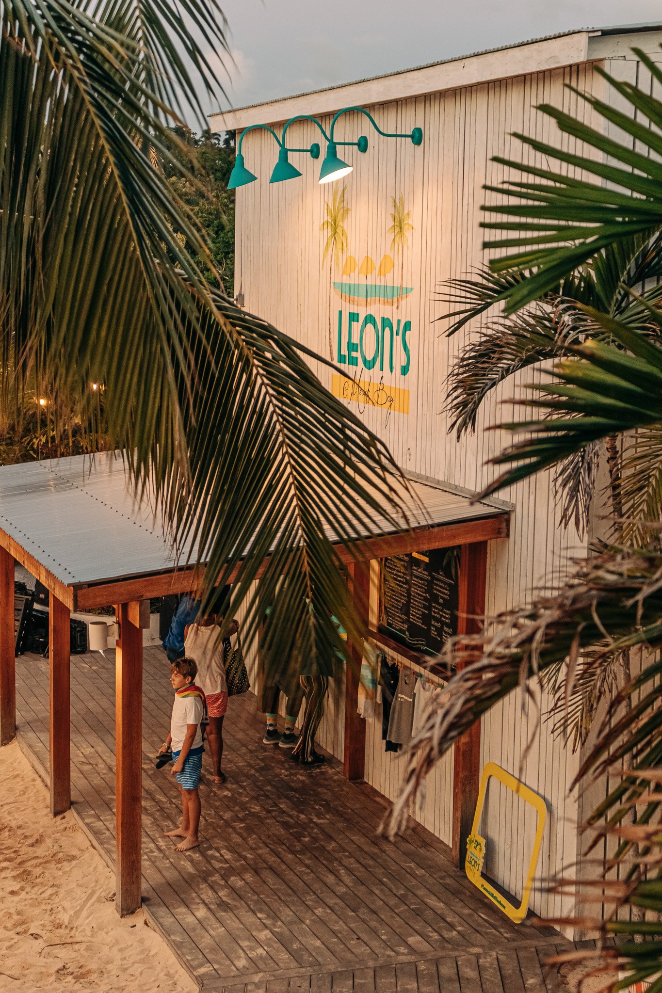 People waiting in line at a wooden food stand with a sign that reads 'Leon's' and a menu board behind it, surrounded by palm trees.