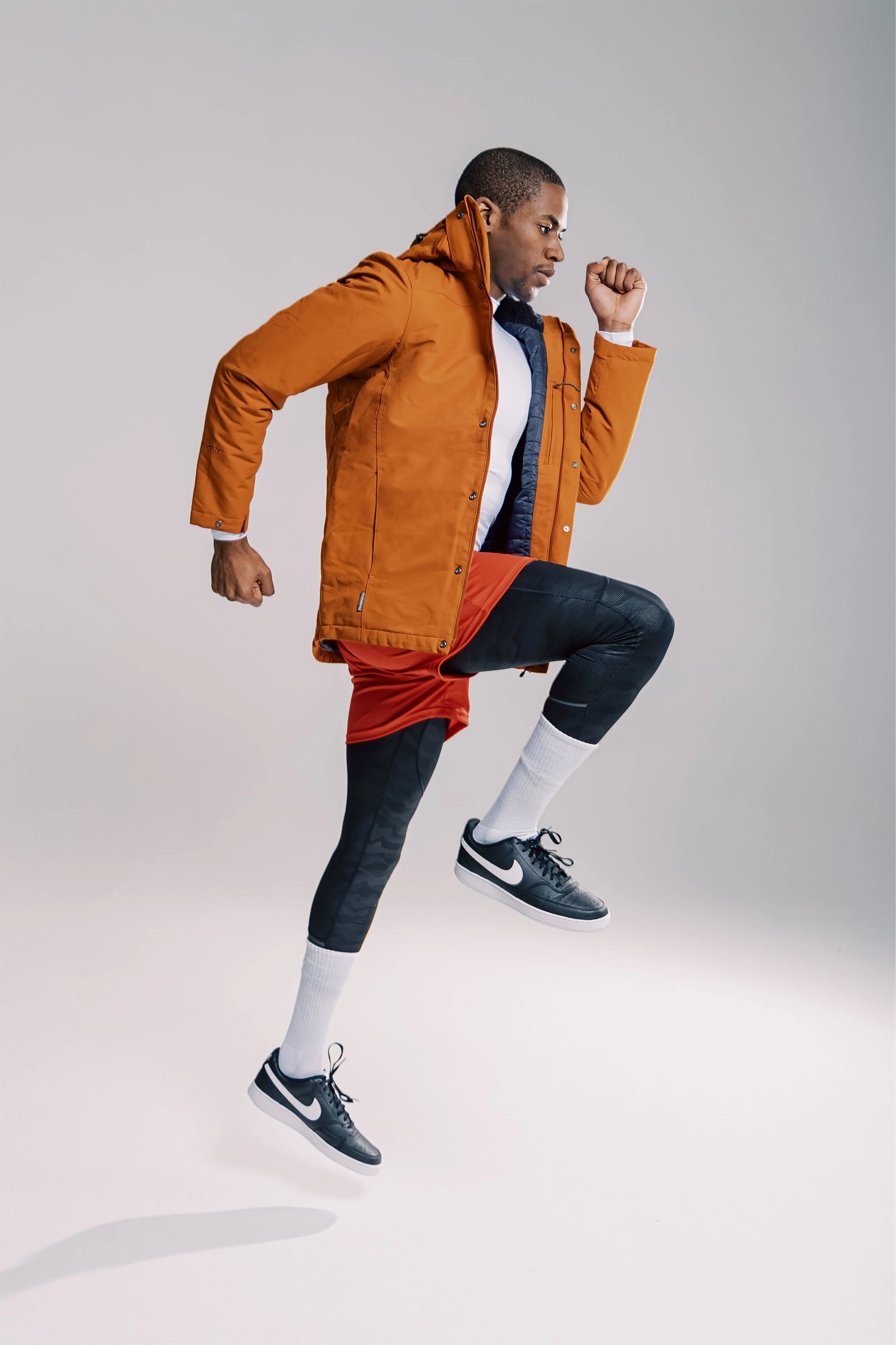 A man in athletic gear mid-air, wearing a brown jacket, red shorts, black leggings, white socks, and black Nike sneakers, posed as if running or jumping.