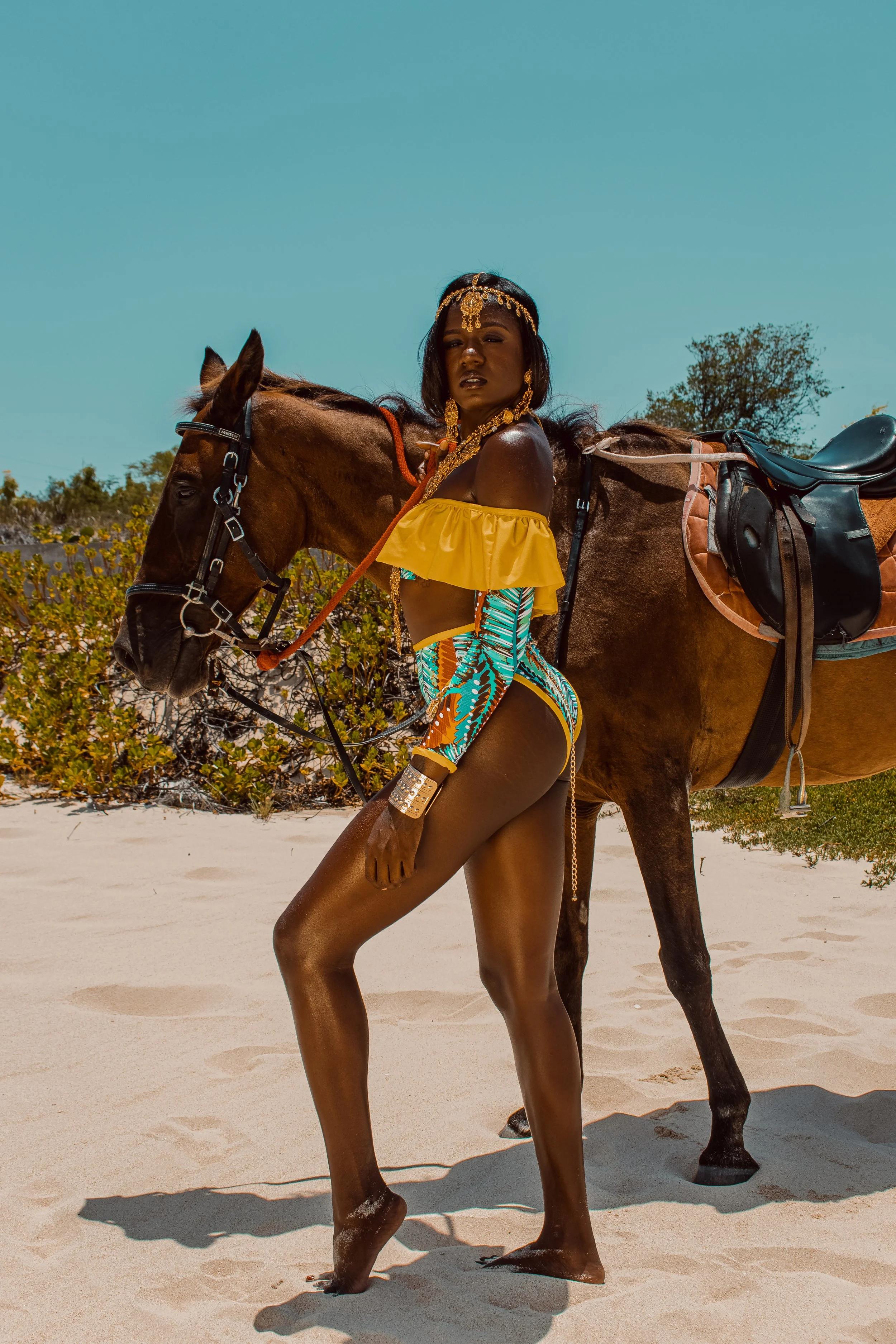 A woman in colorful swimwear and jewelry standing barefoot on a sandy beach, holding a horse's bridle with a saddle and trees in the background.