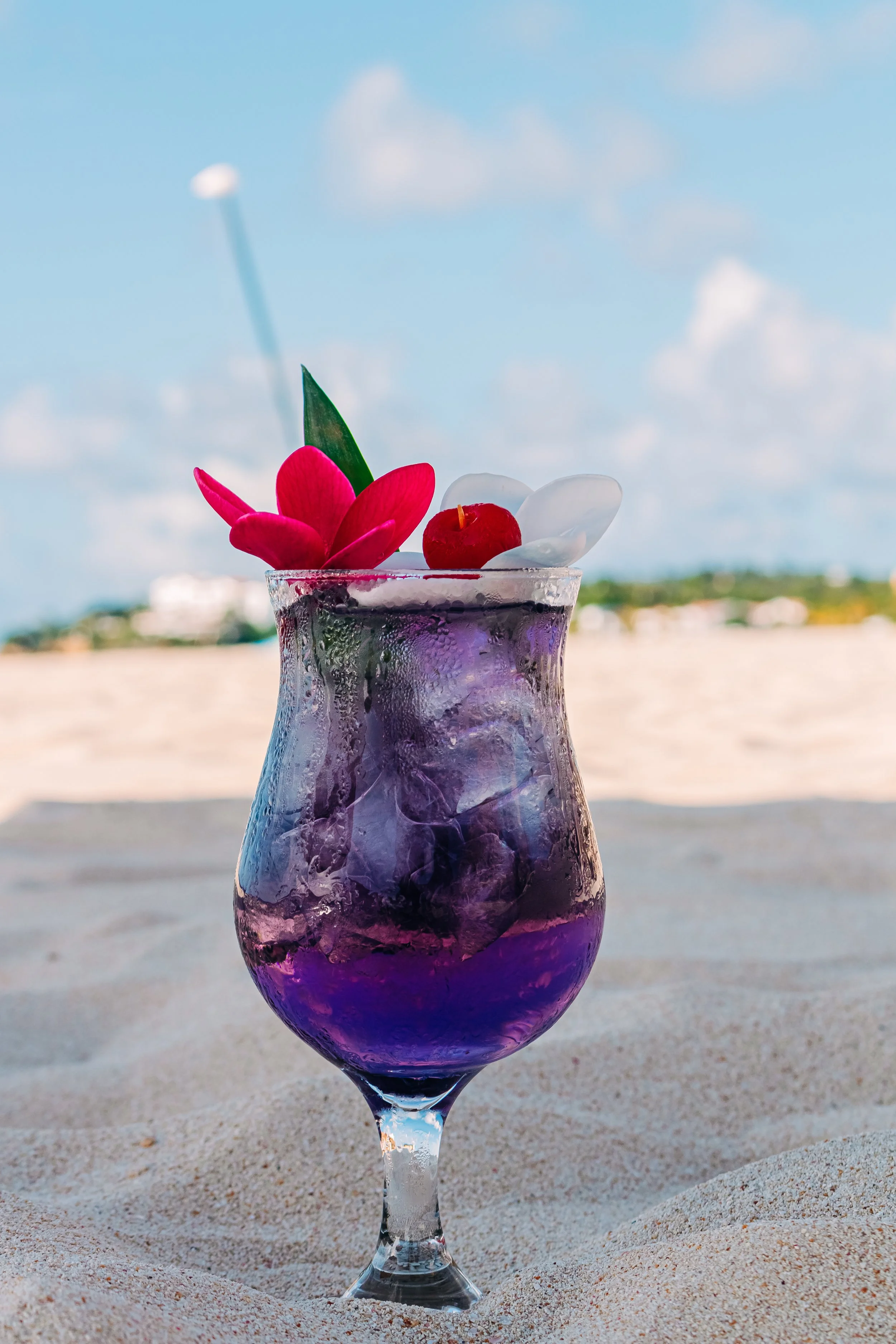 A purple cocktail with ice cubes, a pink flower, a green leaf, and a red cherry, set on sandy beach with a blue sky and clouds in the background.
