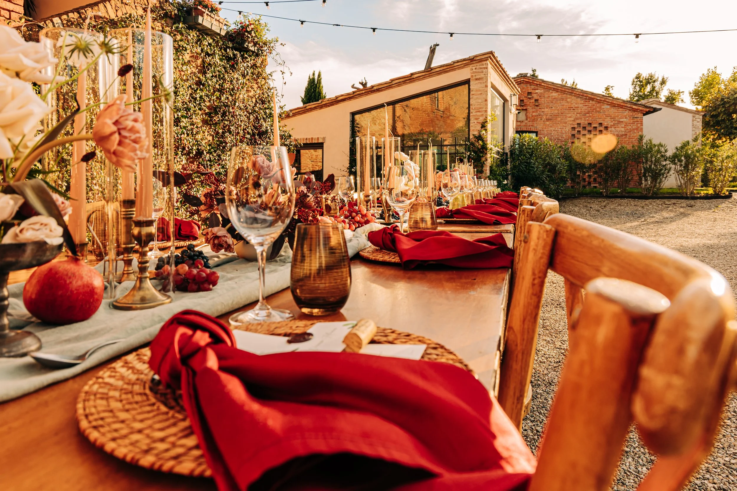 An outdoor dining setup with a long table, red napkins, wine glasses, and a centerpiece with flowers and fruits. Surrounding the table are wooden chairs, and in the background, there are brick buildings and greenery. String lights are hanging above, 