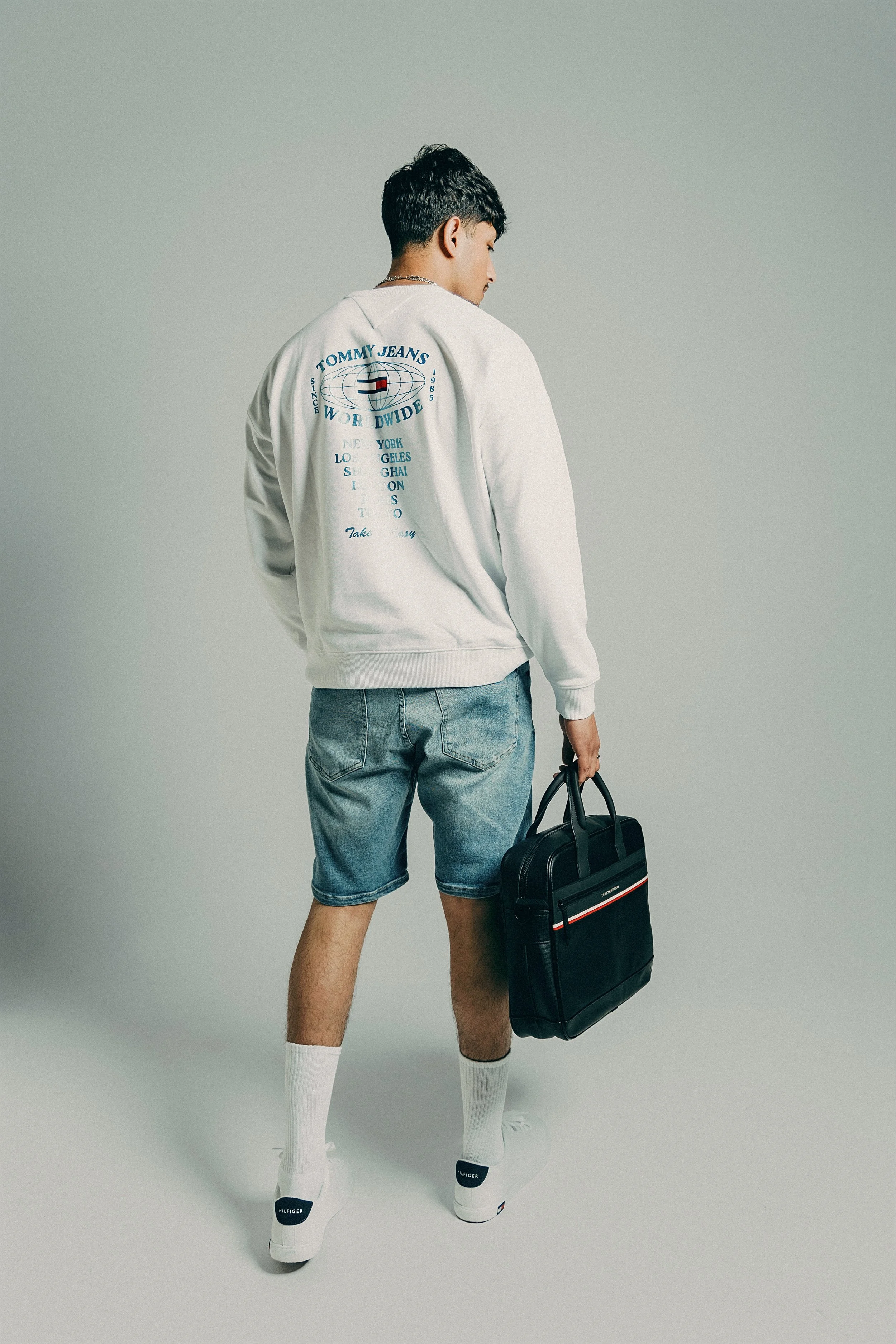 Young man standing with his back to the camera, wearing a white Tommy Jeans sweatshirt, denim shorts, white socks, and sneakers, holding a black briefcase.