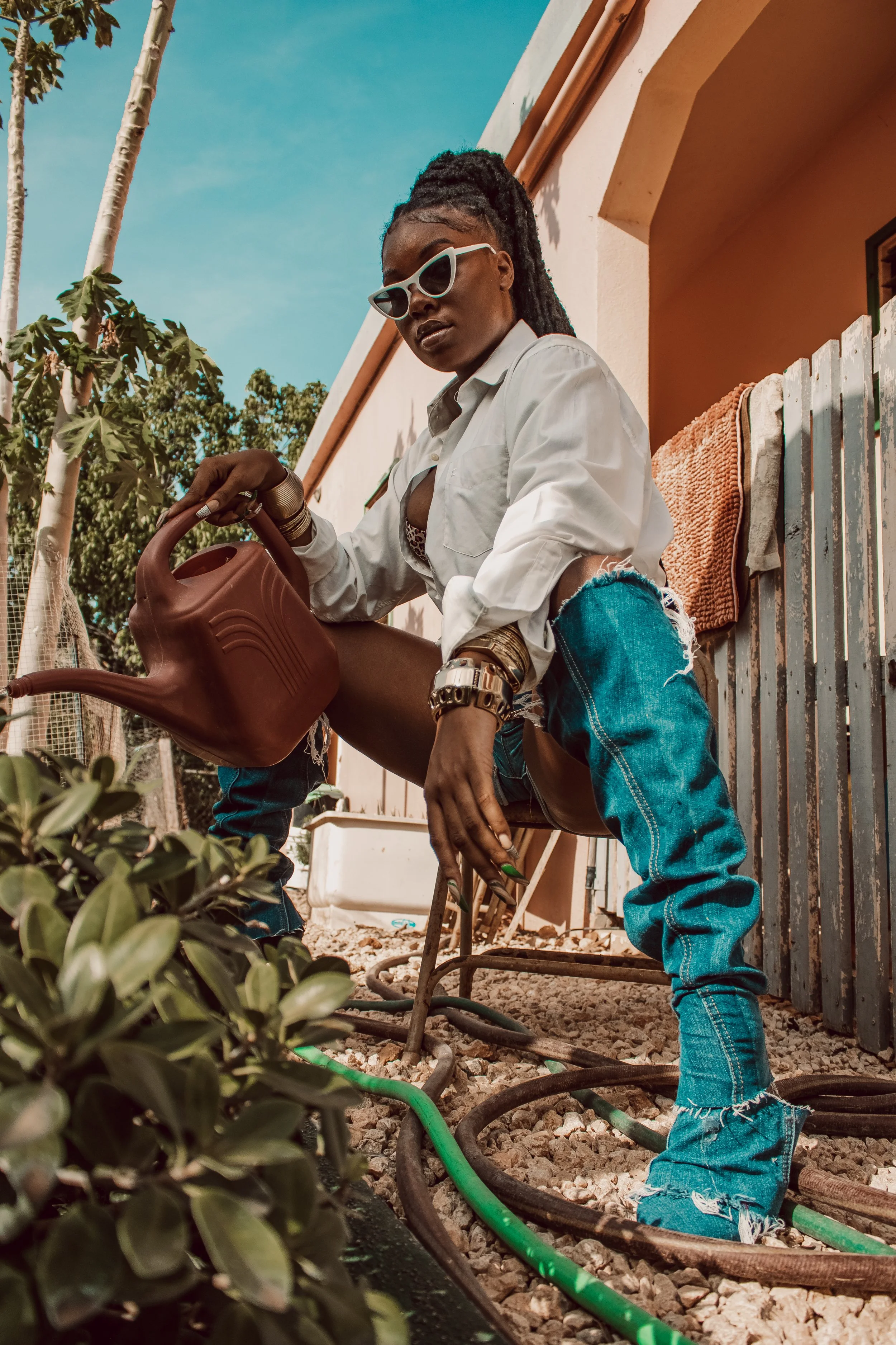 A woman with dark braided hair, sunglasses, and gold jewelry watering plants outside during daytime. She is wearing a white shirt and ripped blue jeans, with a garden hose and watering can, in front of a pink building and tree.