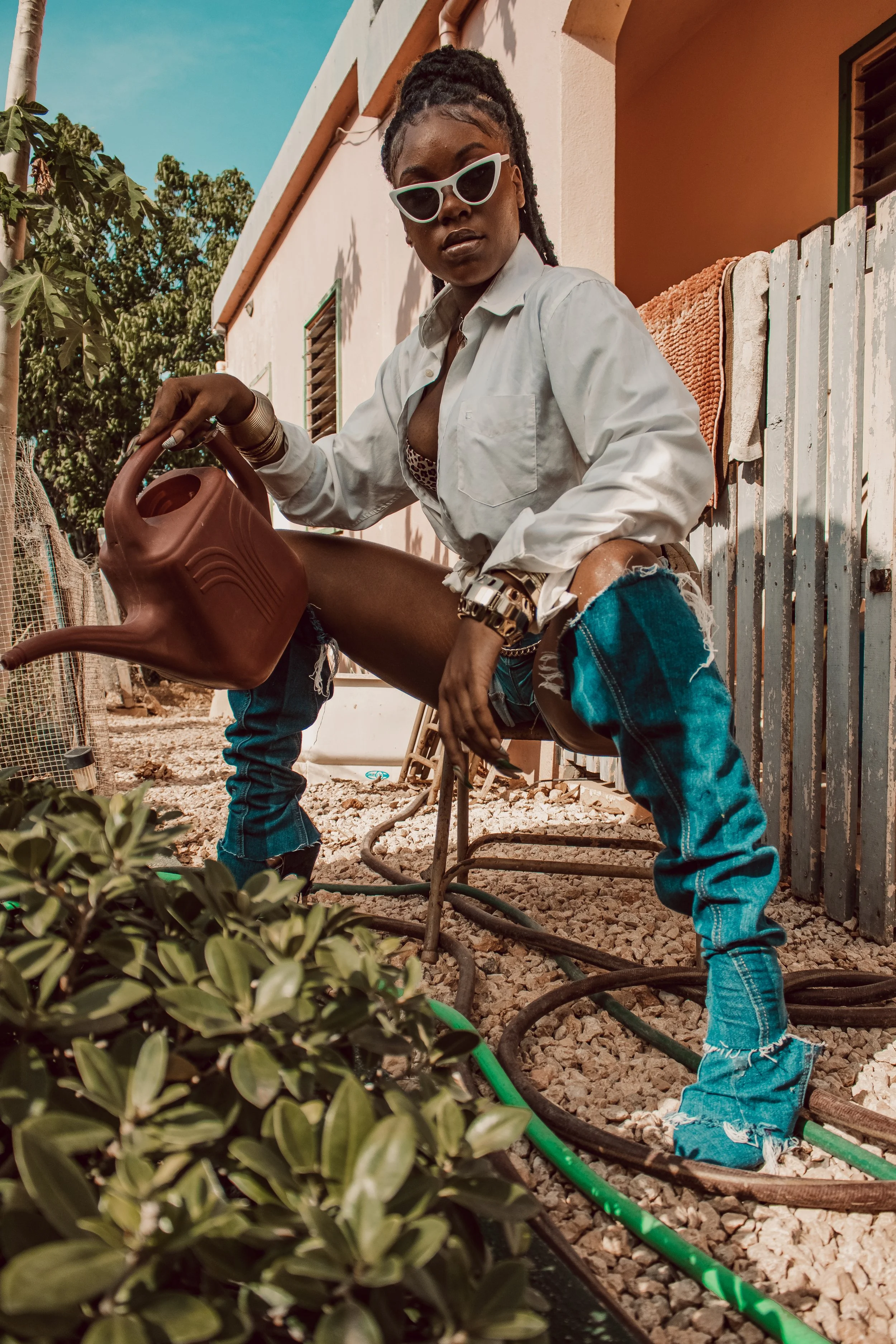 A woman with sunglasses, wearing a white shirt and ripped jeans, watering plants outside a building.