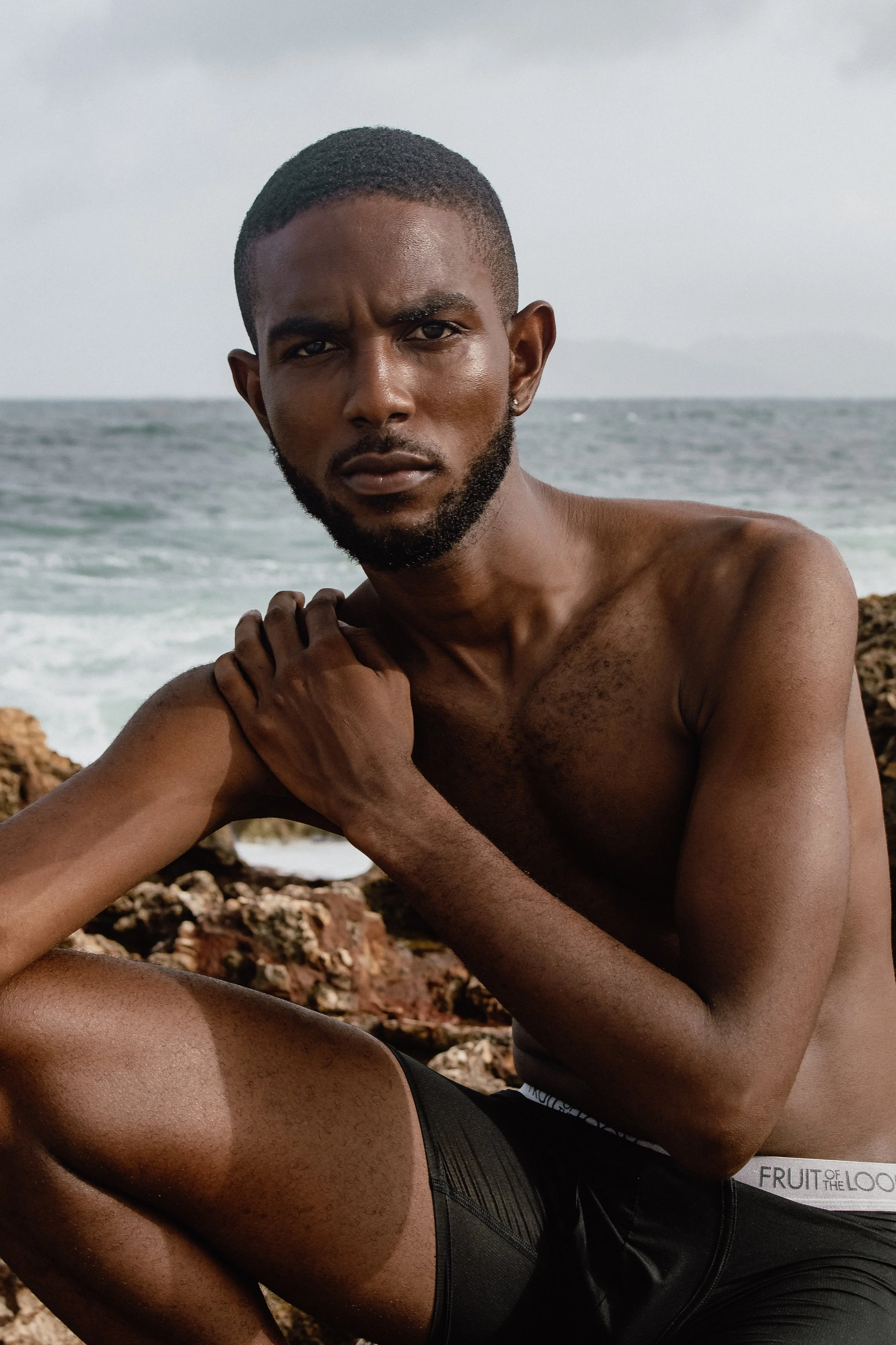 A shirtless man with a beard on a rocky beach, looking directly at the camera, with the ocean and cloudy sky in the background.