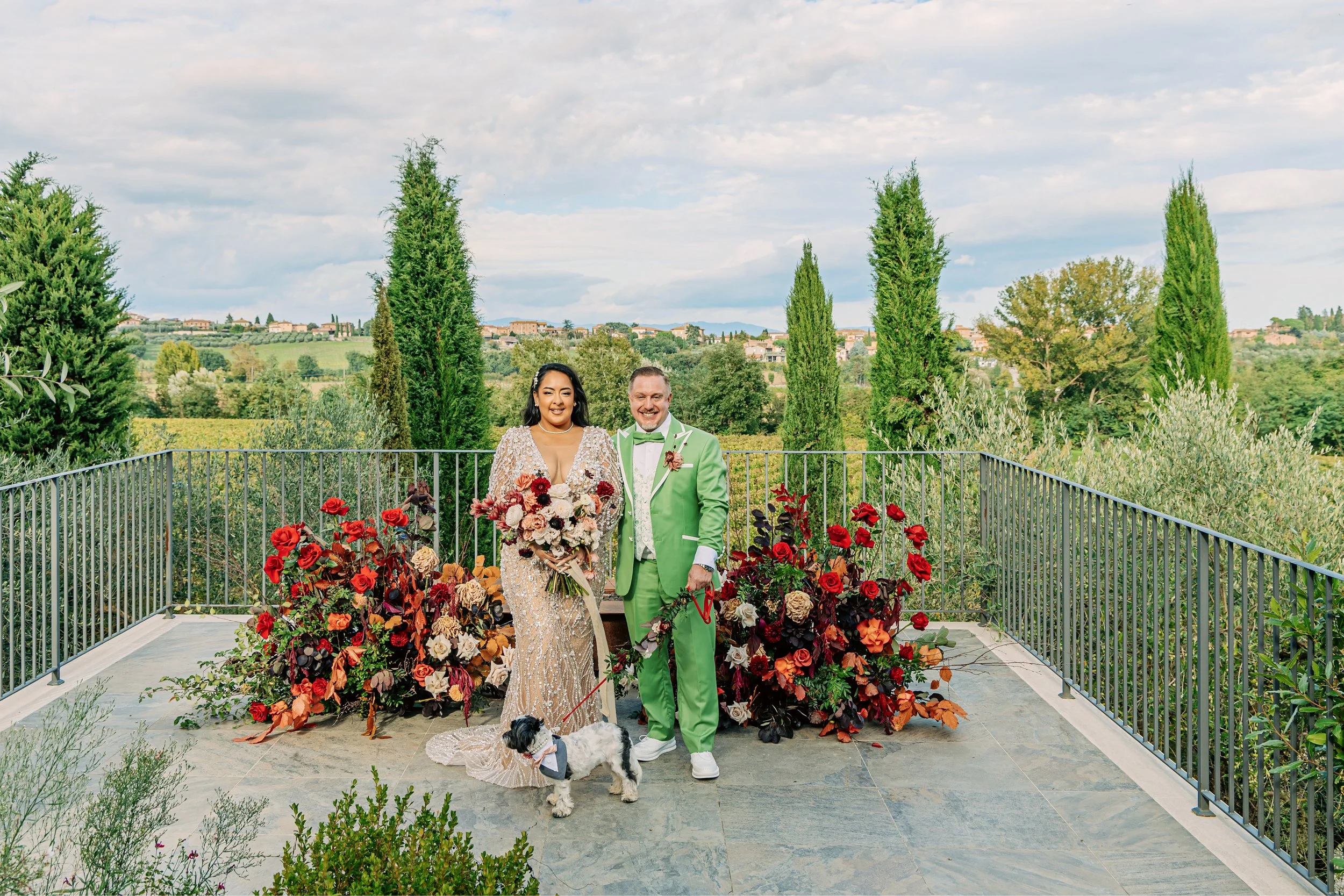 A bride and groom standing together on a balcony decorated with red and pink flowers, with a scenic landscape of trees and rolling hills in the background. The bride wears a sparkly dress and holds a bouquet, while the groom wears a green suit. A sma