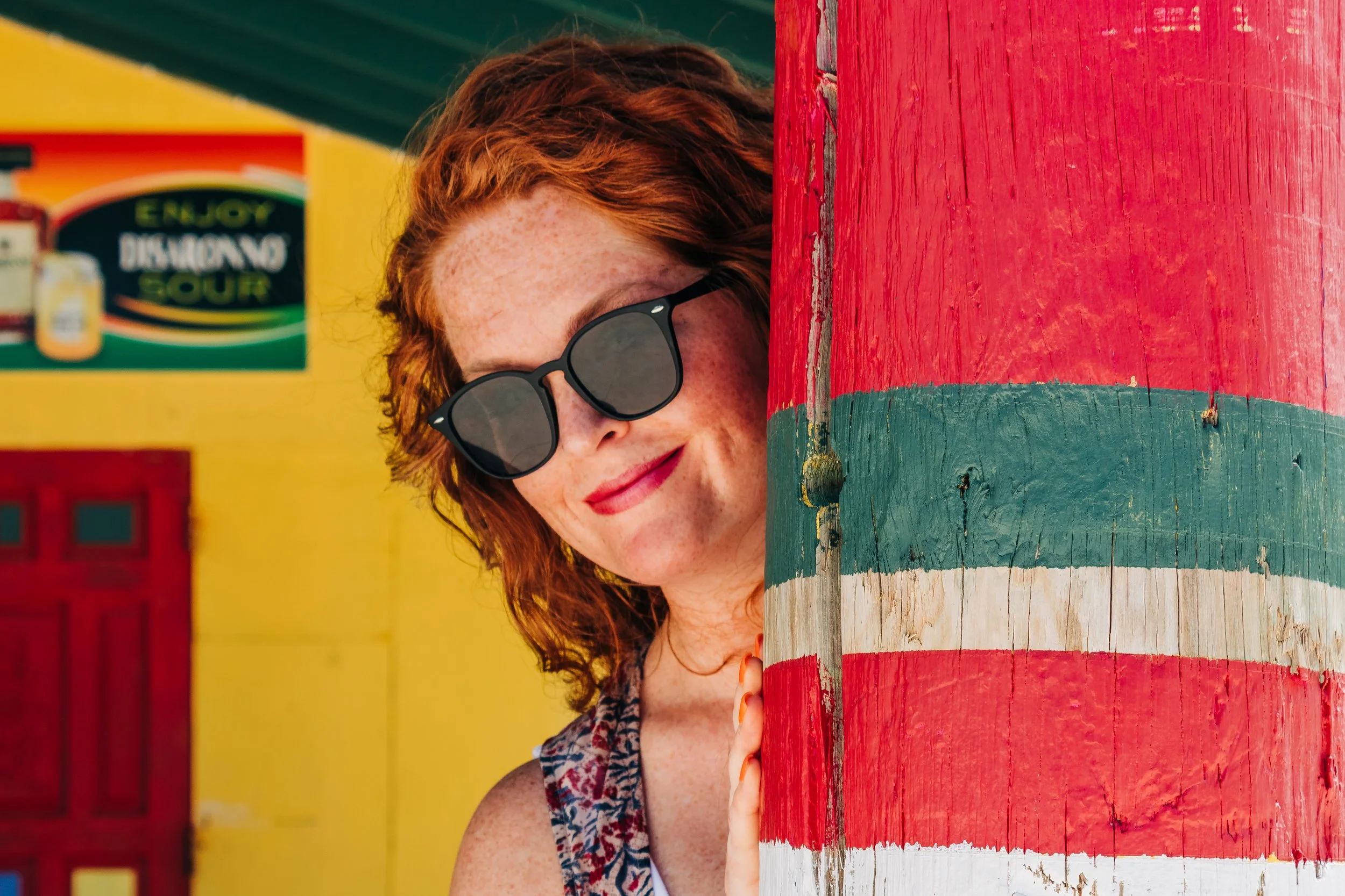 A woman with red hair and sunglasses peeks from behind a colorful red, green, and white painted wooden pole.
