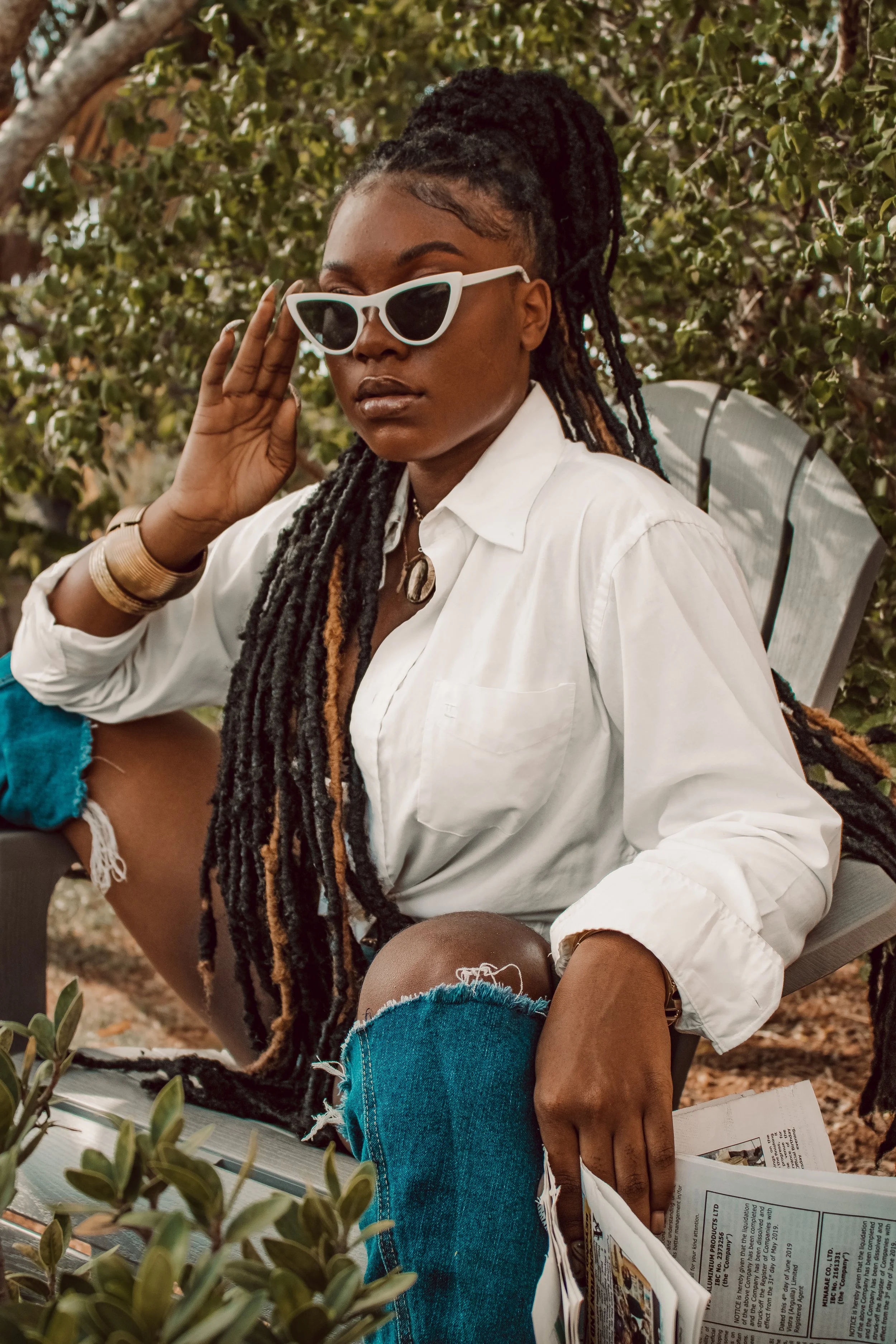 A woman with long dreadlocks, wearing sunglasses and a white shirt, sits outdoors on a wooden chair among green bushes. She holds a newspaper in one hand and touches her face with the other, displaying a confident pose.