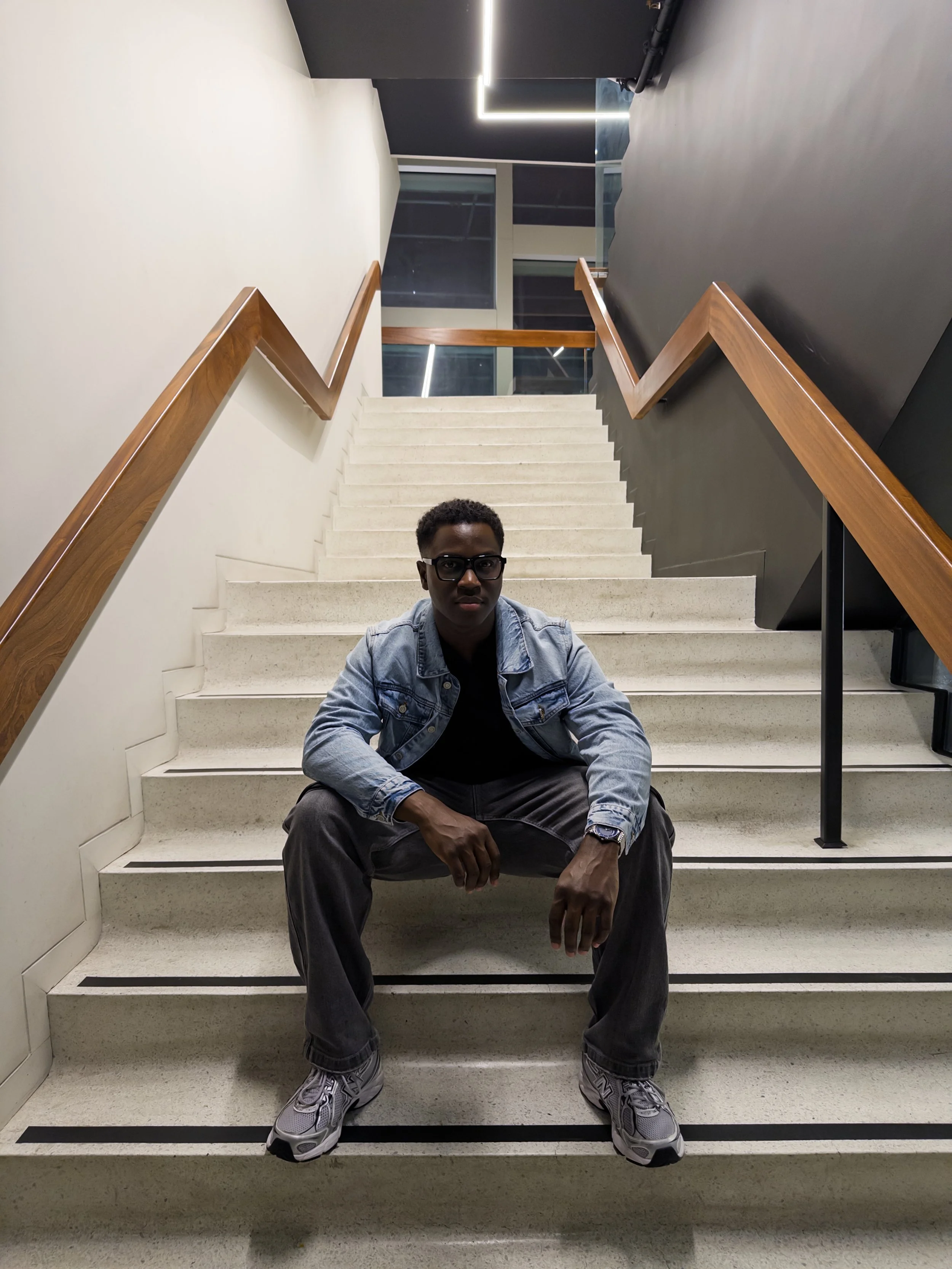 Jonathan Gumbs also known as J.Austin Pro (JAPro) with glasses in denim jacket sitting on staircase in modern building, looking at camera.