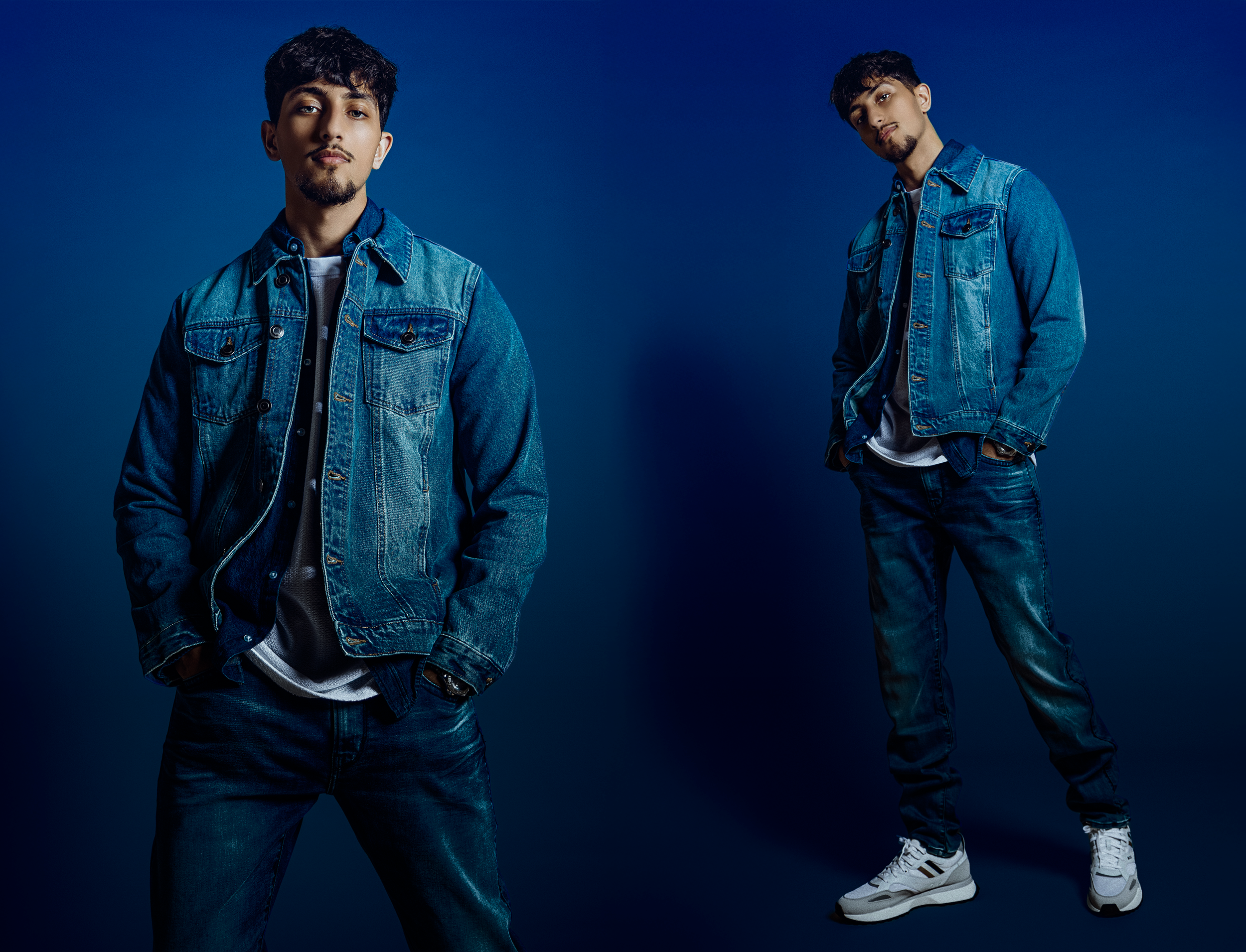 Two young men in denim jackets and jeans, standing against a blue background, posing for a photo.