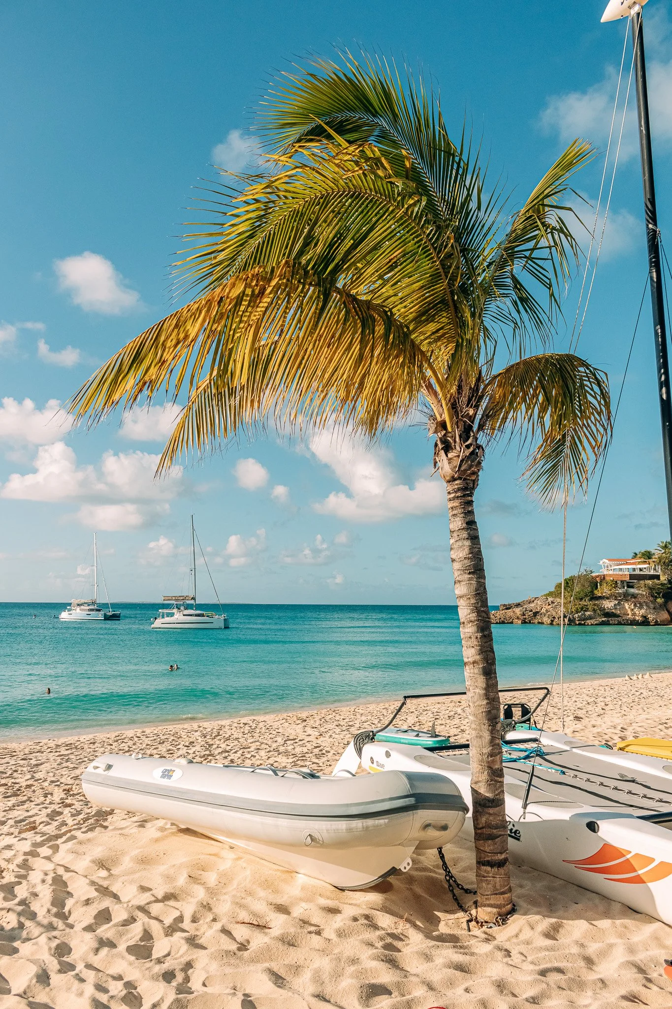A tropical beach scene with inflatable boats on the sandy shore, a palm tree, and sailboats in the turquoise water under a blue sky with scattered clouds.