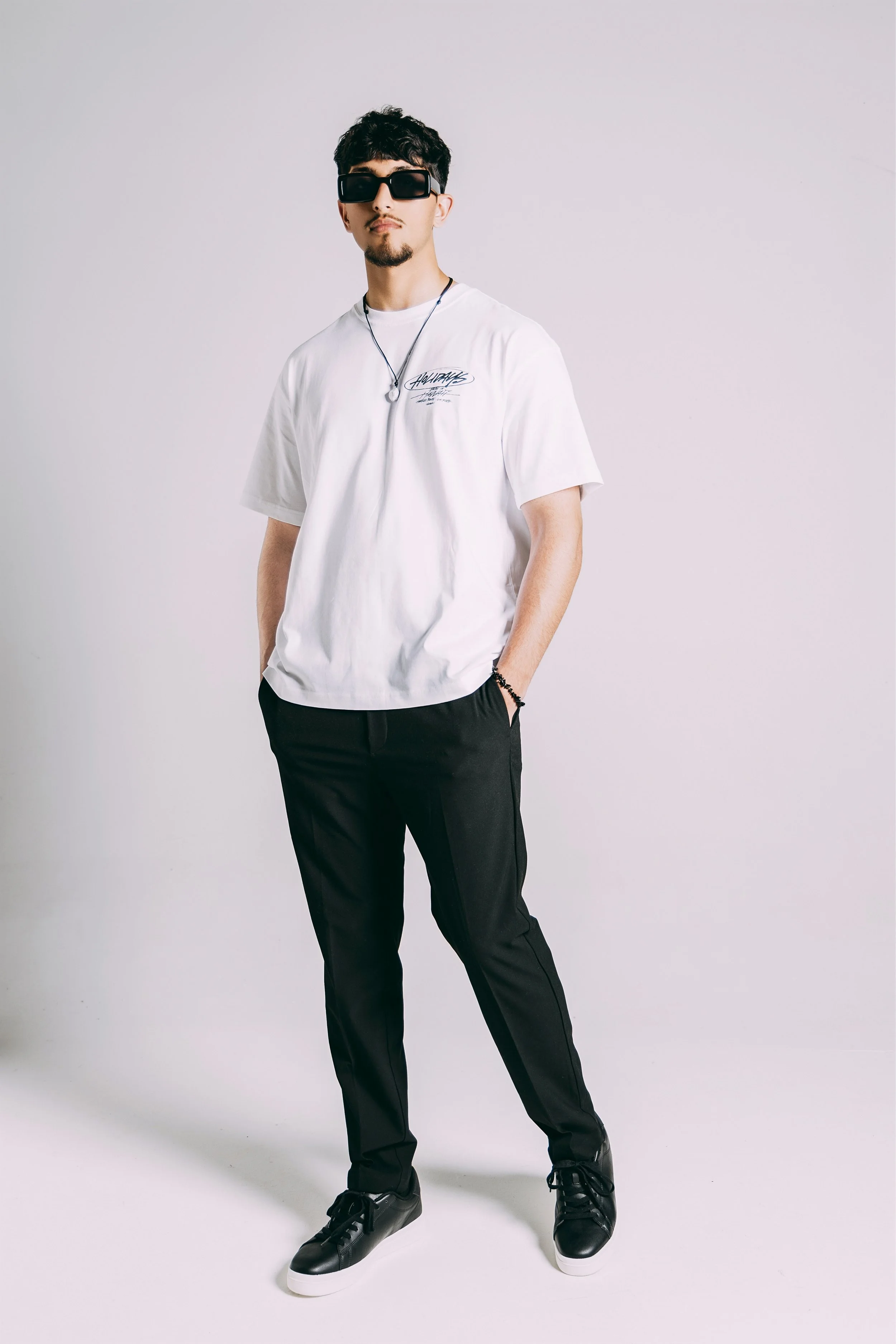 A young man standing against a plain white background, wearing black sunglasses, a white oversized t-shirt with text, black pants, black sneakers, and a black beaded bracelet.