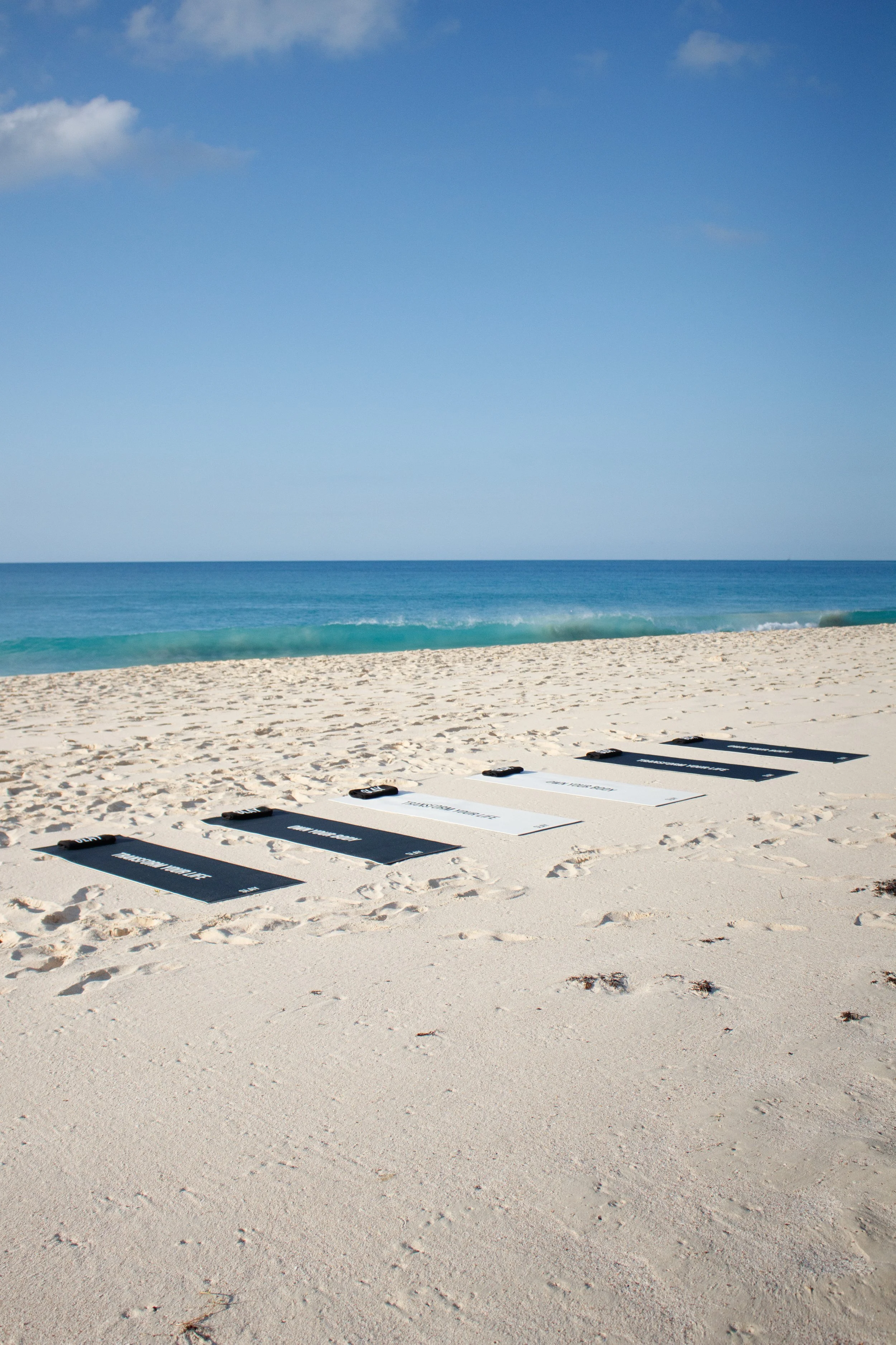 Six black and white yoga mats with black straps placed on the sandy beach near the shoreline, with clear blue ocean water and sky in the background.
