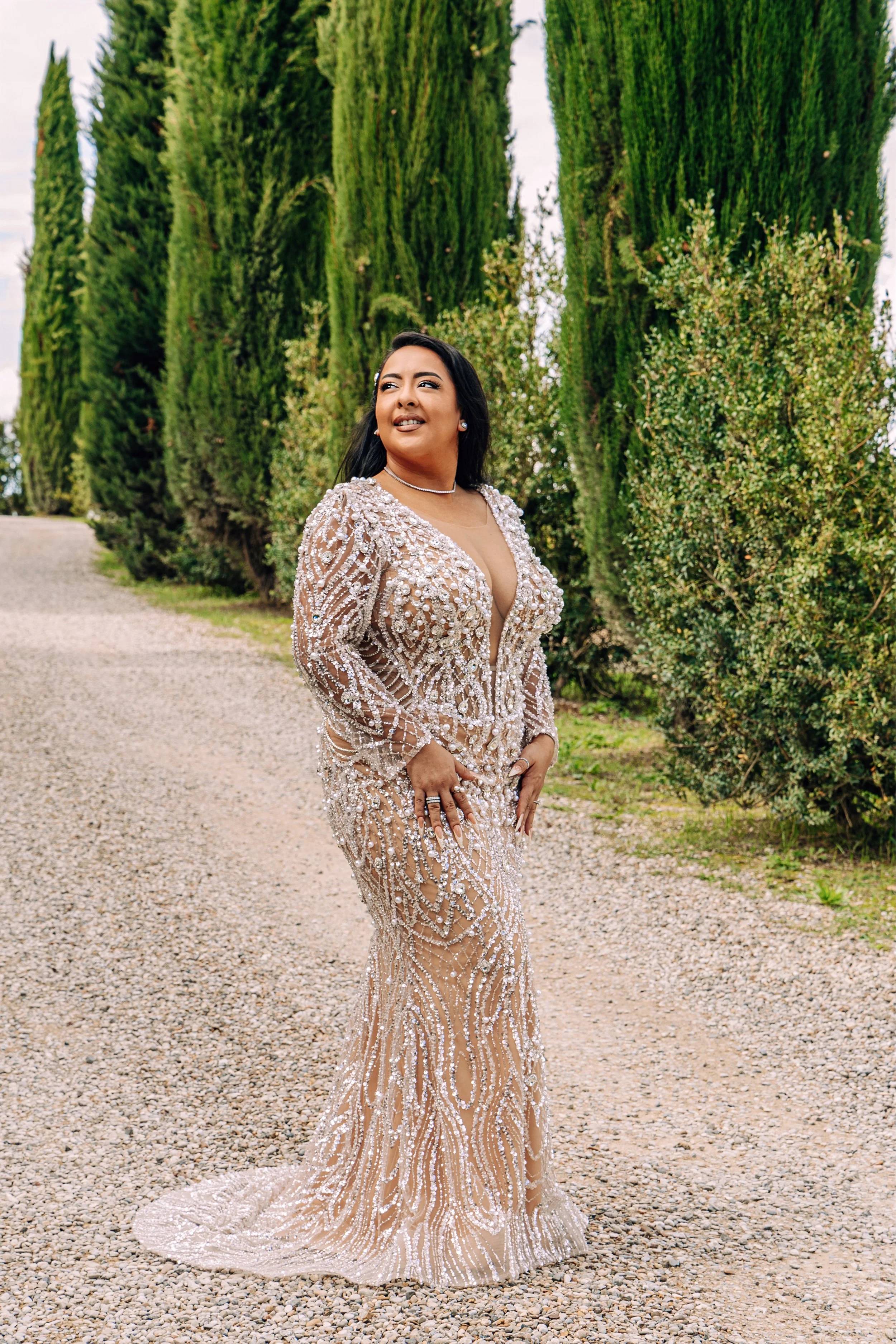 A woman in a sparkling, beaded gown stands on a gravel path with tall, green cypress trees in the background.