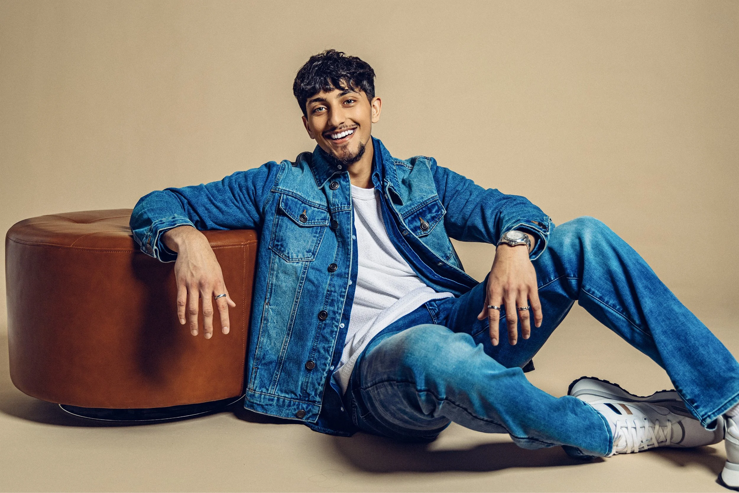 Young man sitting on the floor leaning against a brown ottoman, wearing a denim jacket and jeans, white sneakers, smiling, with beige background.
