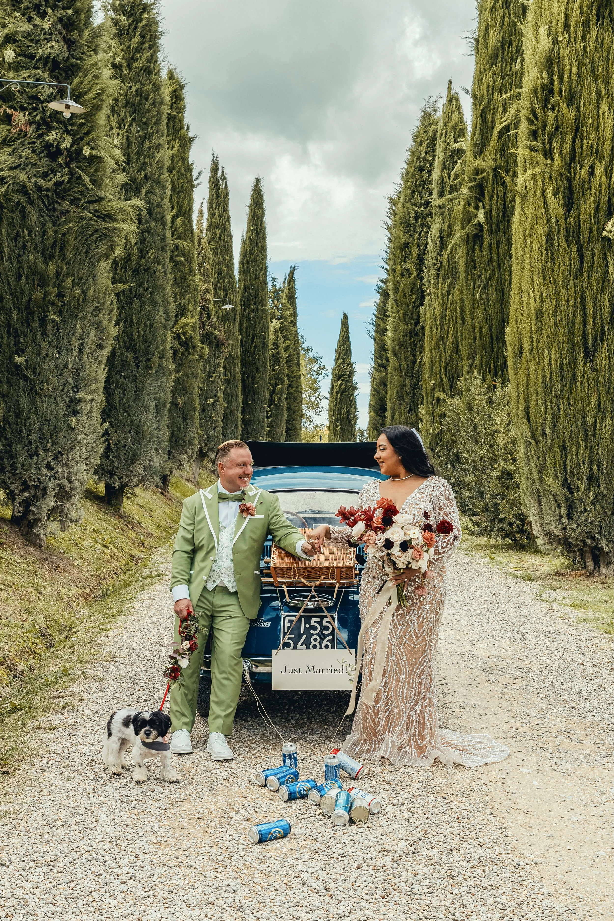 A newly married couple walks away from a vintage car with a sign that says 'Just Married' on a gravel path surrounded by tall greenery. The groom is dressed in a light green suit with a boutonniere, and the bride is in a sparkling, beaded wedding gow
