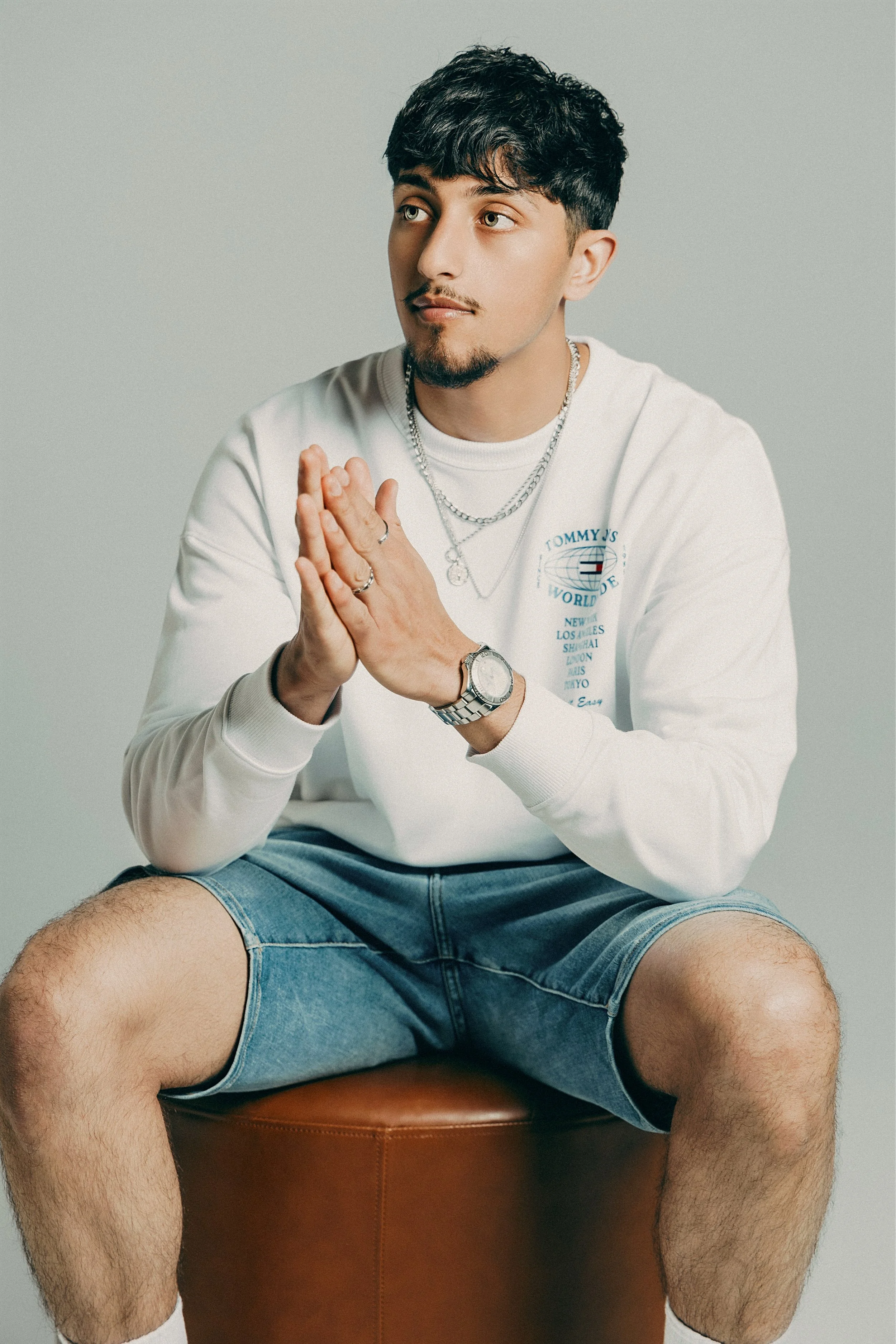 A young man with dark hair and facial hair, sitting on a brown stool against a gray background. He is wearing a white sweatshirt, denim shorts, multiple necklaces, a watch, and rings. His hands are pressed together in front of his chest, and he is lo