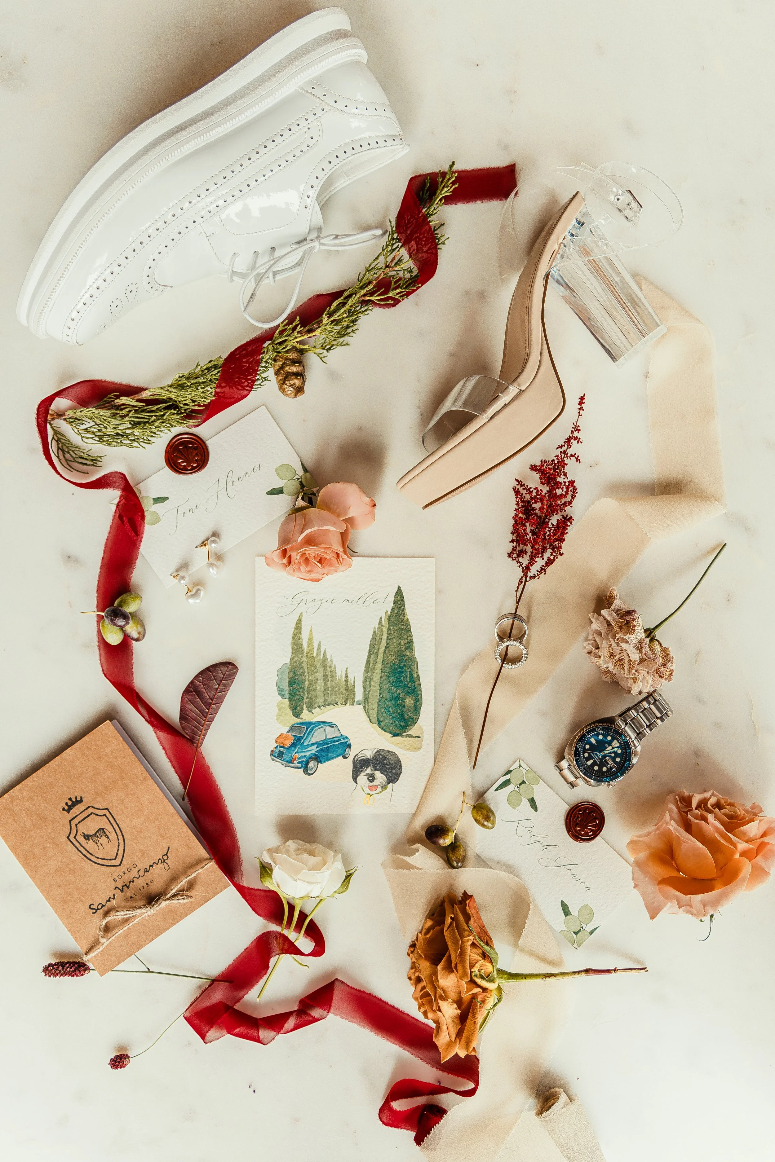 A flat lay arrangement with flowers, ribbons, postcards, a watch, a small box, and decorative objects on a light surface.