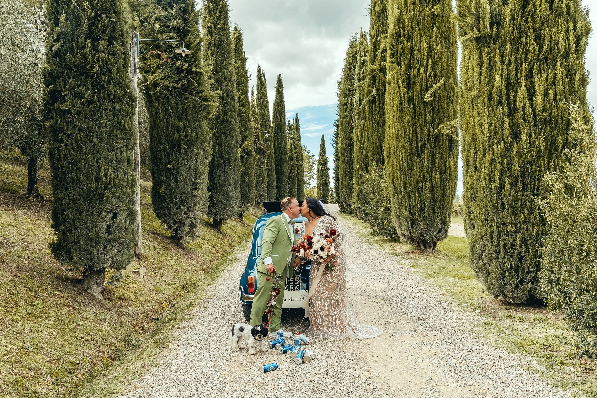 A newly married couple dressed in formal attire sharing a kiss outdoors next to a vintage blue car with a 'Just Married' sign. The bride wears a white lace wedding gown and holds a bouquet, while the groom wears a light green suit. They are standing 