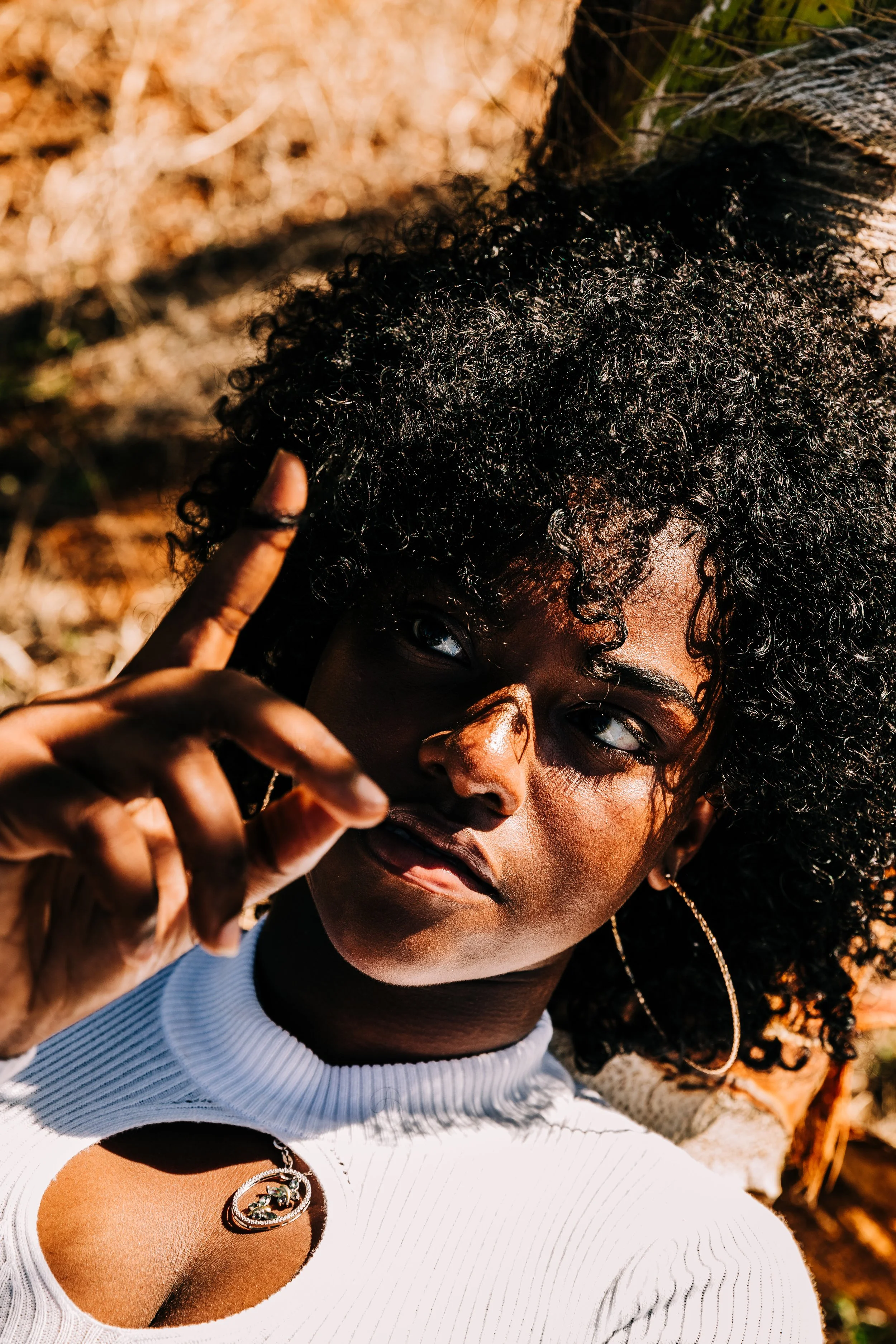 A woman with dark curly hair, wearing a white top with a small cutout, gold hoop earrings, a necklace with a circular pendant, and making a peace sign with her hand, outdoors during sunset.