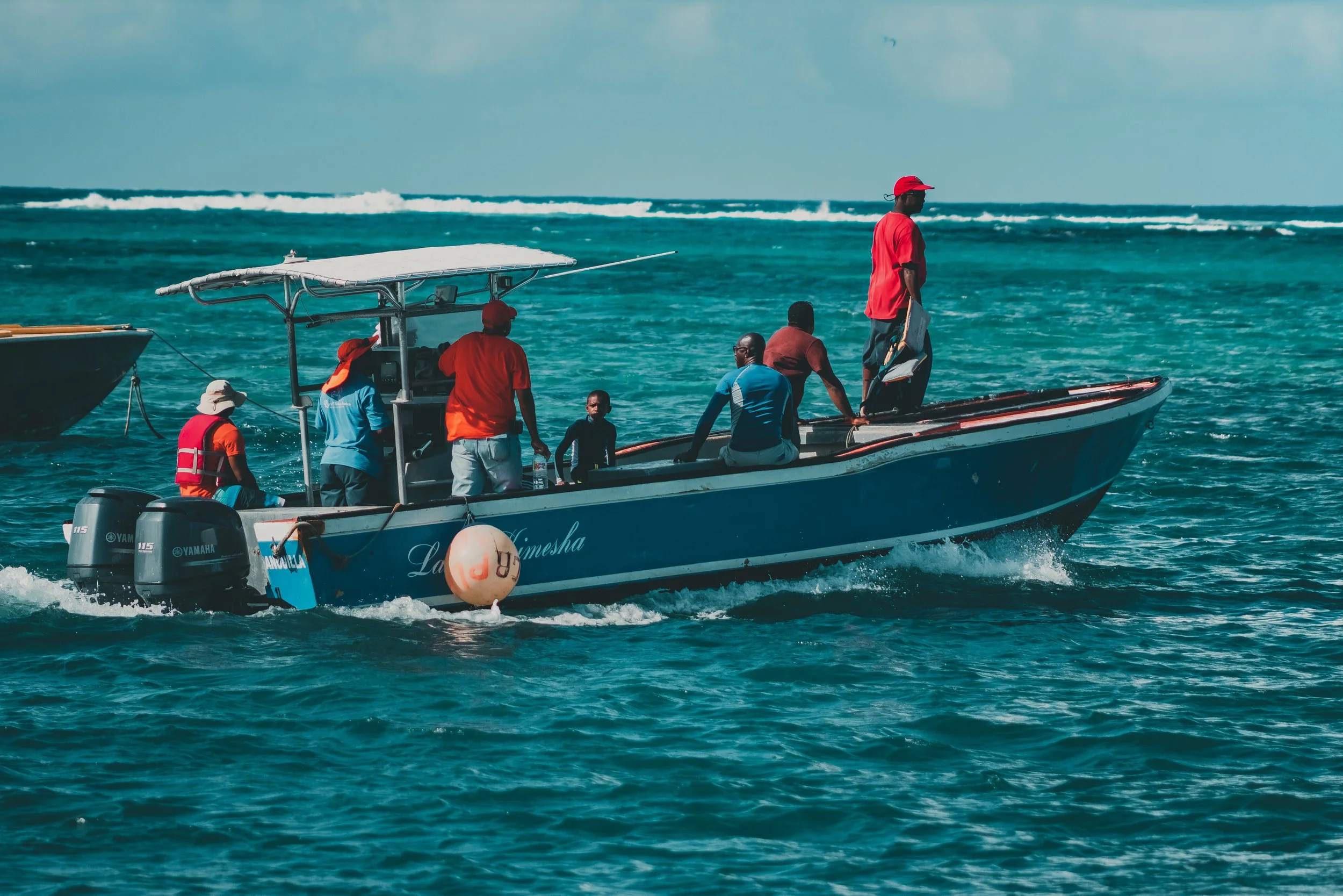 People riding on a motorboat on the ocean, some wearing red shirts and hats, others in blue, with a buoy floating nearby and waves in the background.