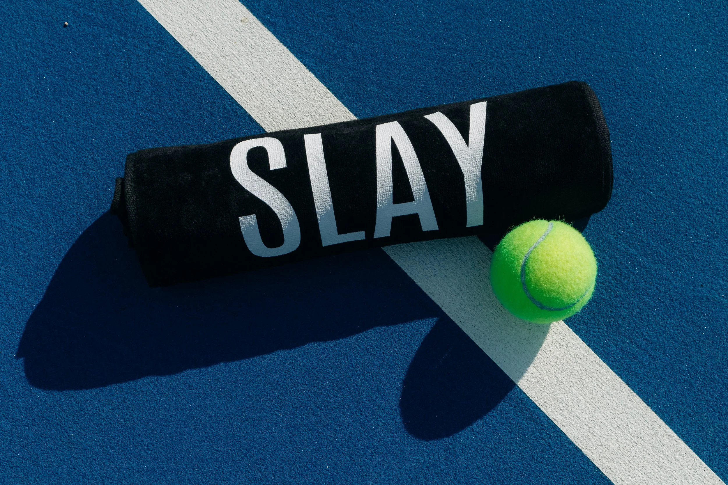 A rolled up black towel with the word 'STAY' printed in white on it, lying on a blue tennis court with a white line, alongside a bright green tennis ball.