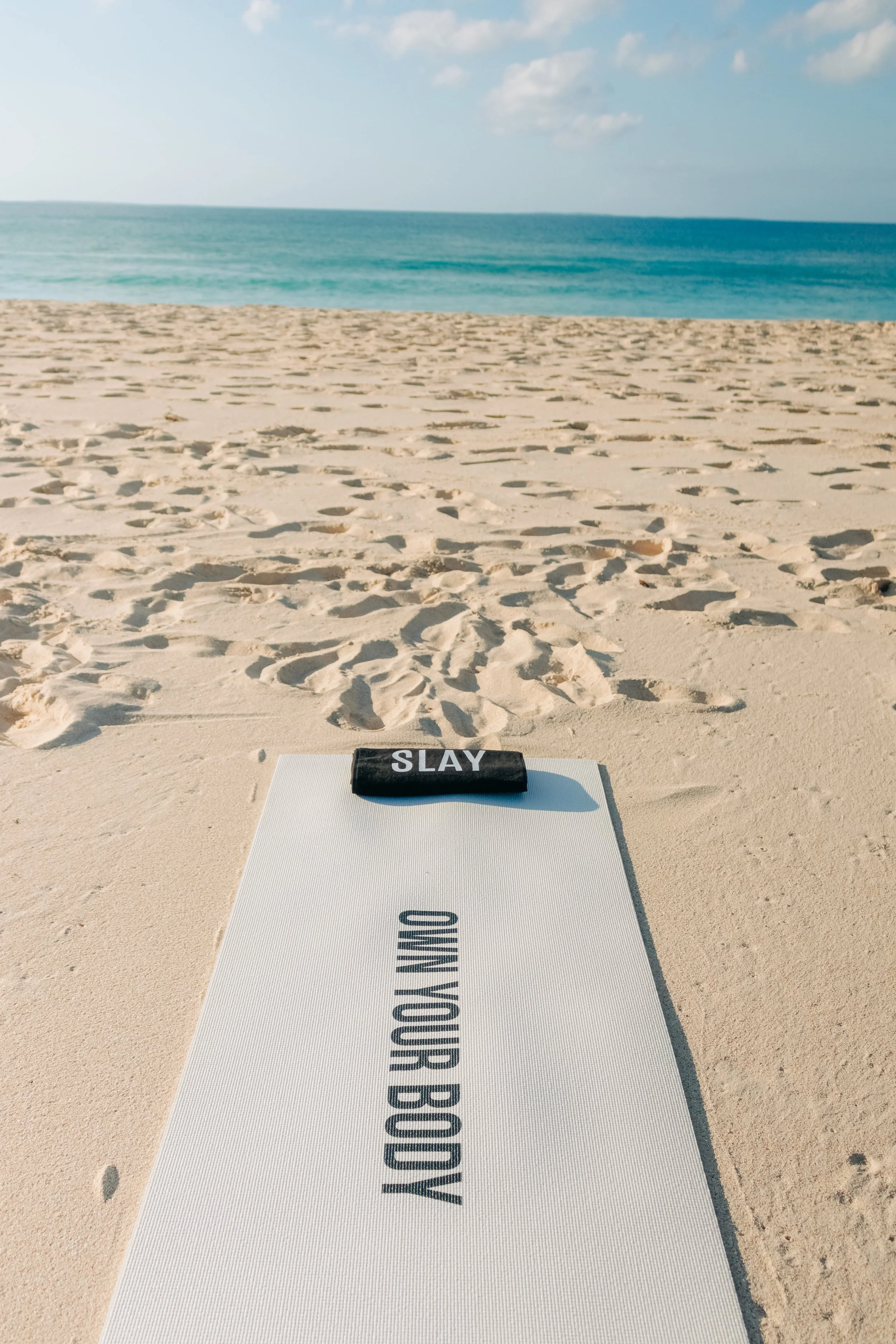 A yoga mat on a sandy beach with the words "RUN YOUR BODY" on it, and a black cloth with "SLAY" on top of the mat. The ocean and blue sky are in the background.