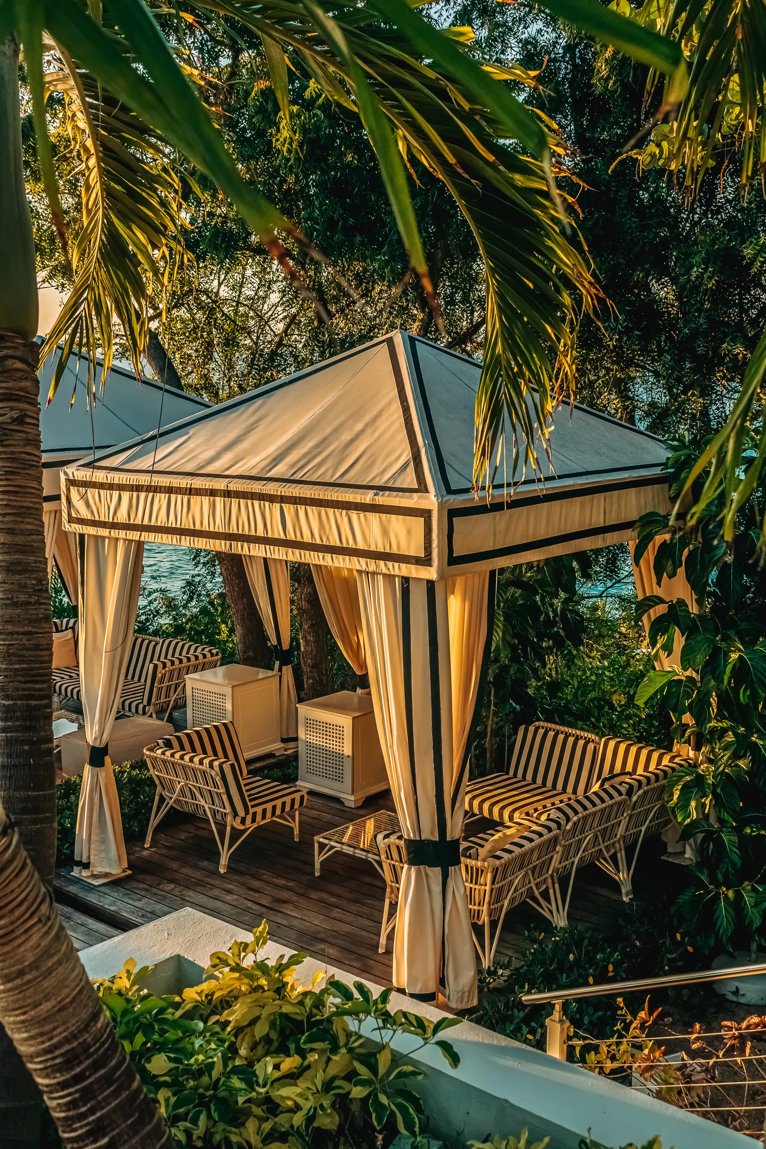 A luxurious outdoor seating area under a canopy with striped cushioned chairs and a cabana with curtains, surrounded by lush green foliage and tropical plants.