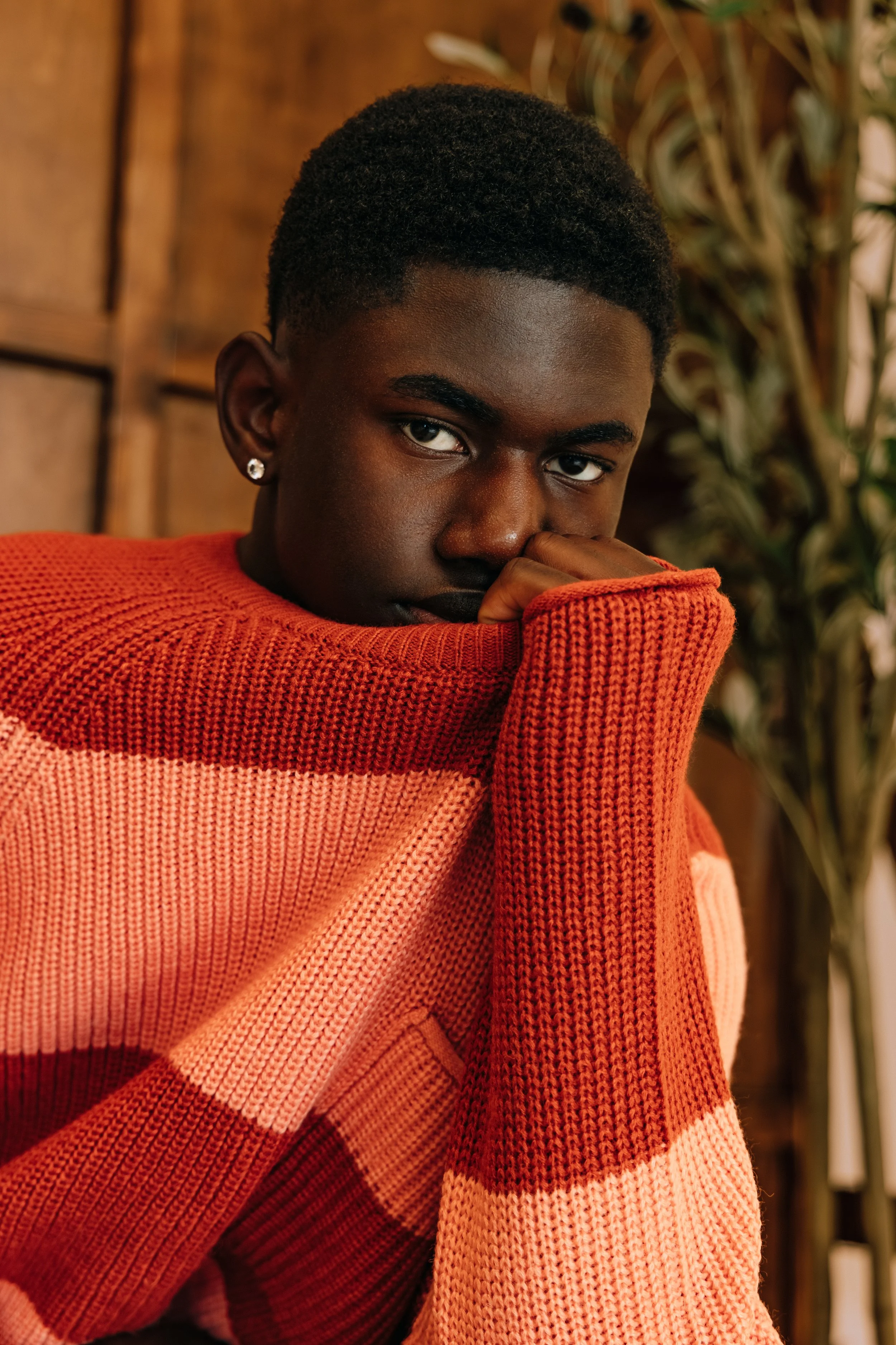 Portrait of a young Black man wearing an orange and pink knit sweater, with short curly hair, looking directly at the camera with his hand covering part of his face. Background includes a wooden wall and green plant leaves.