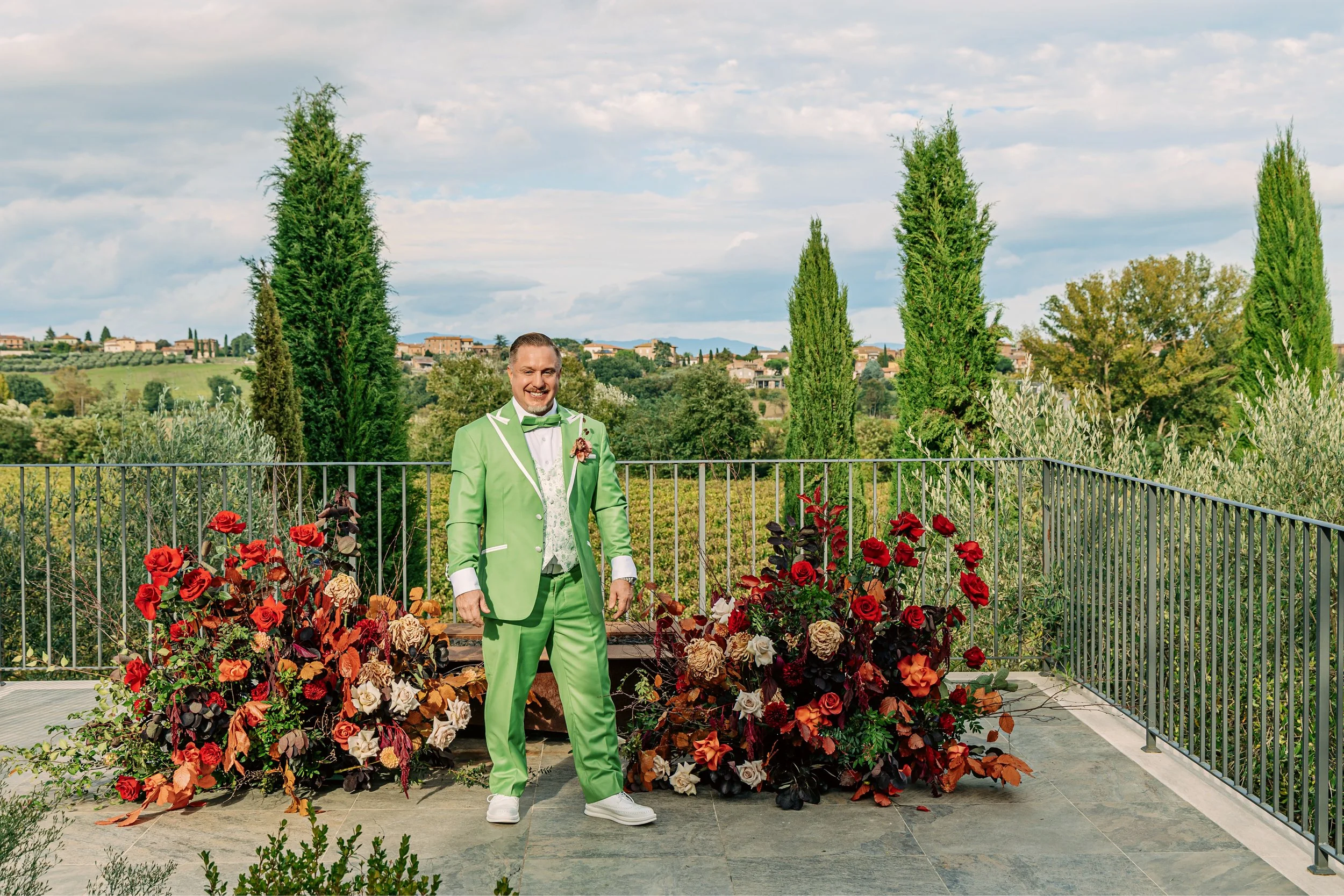 A man in a bright green suit with white accents and a green bowtie, standing in front of a pot of red and orange roses on a balcony with a scenic countryside background, under a partly cloudy sky.