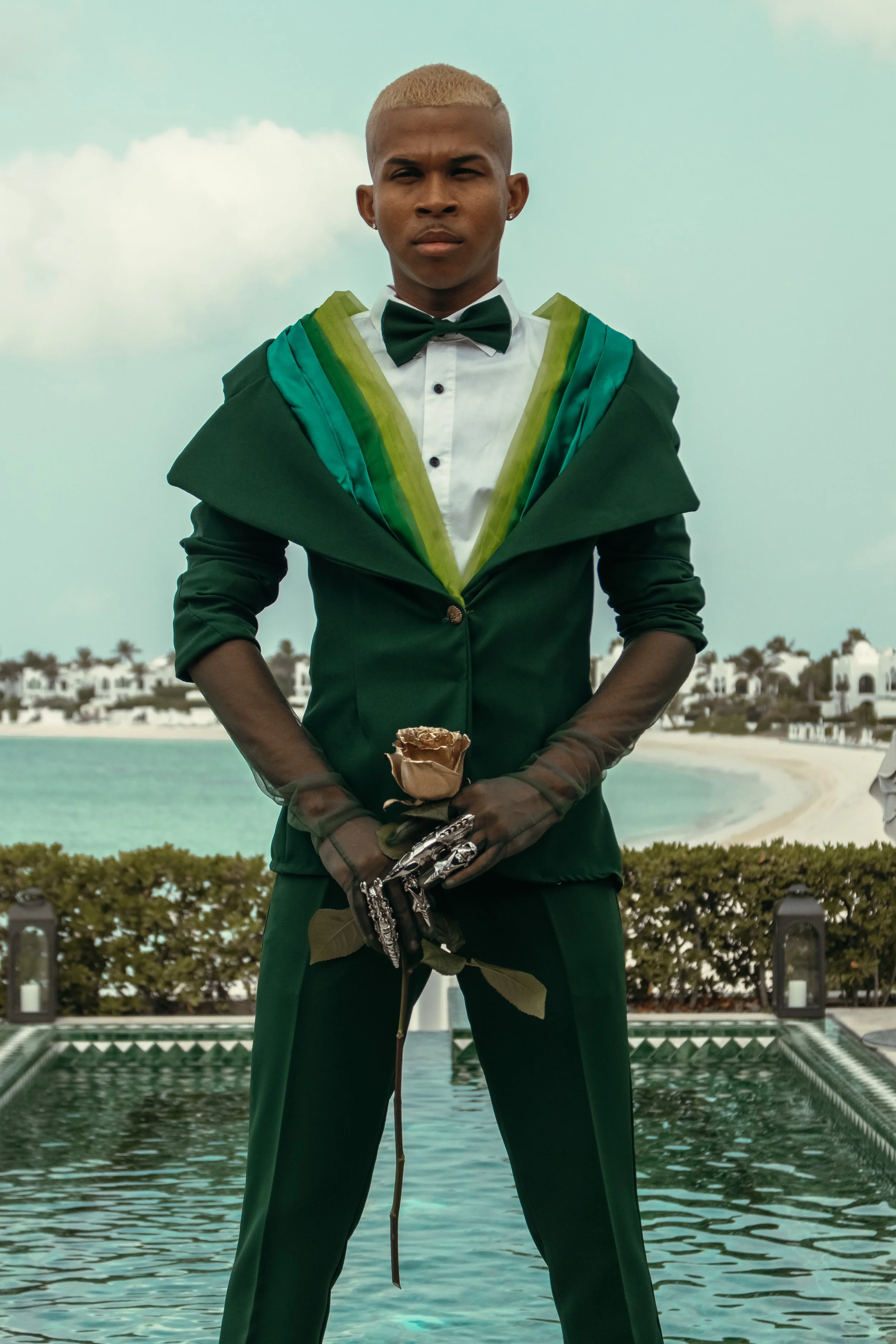 A person standing outdoors near a pool, holding a rose, dressed in a green tuxedo with a white shirt and a green bow tie, with a serious expression.