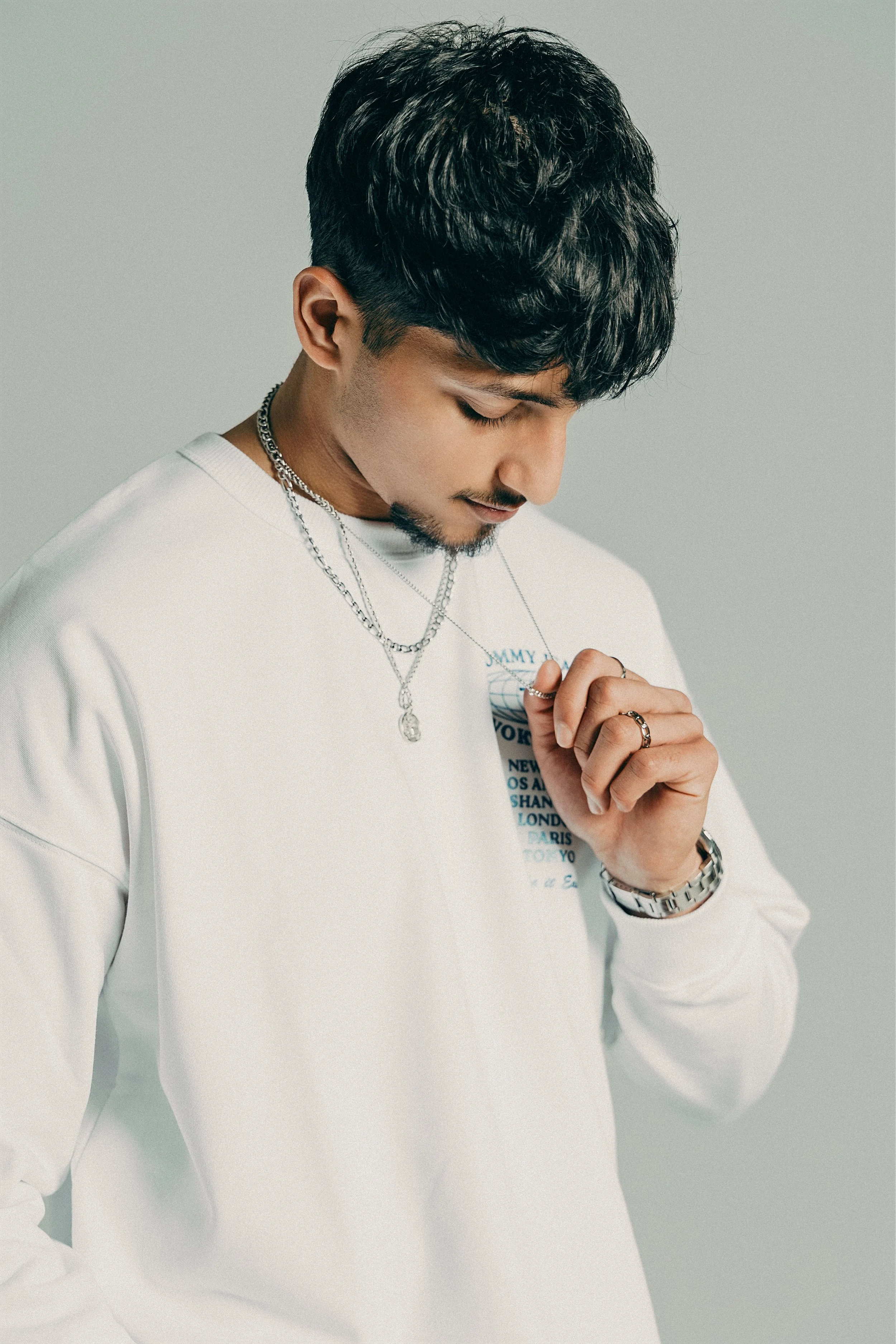 A young man with dark, wavy hair, wearing a white sweatshirt, silver necklaces, a silver wristwatch, and rings, standing with his head tilted downward and hand raised to his chest.