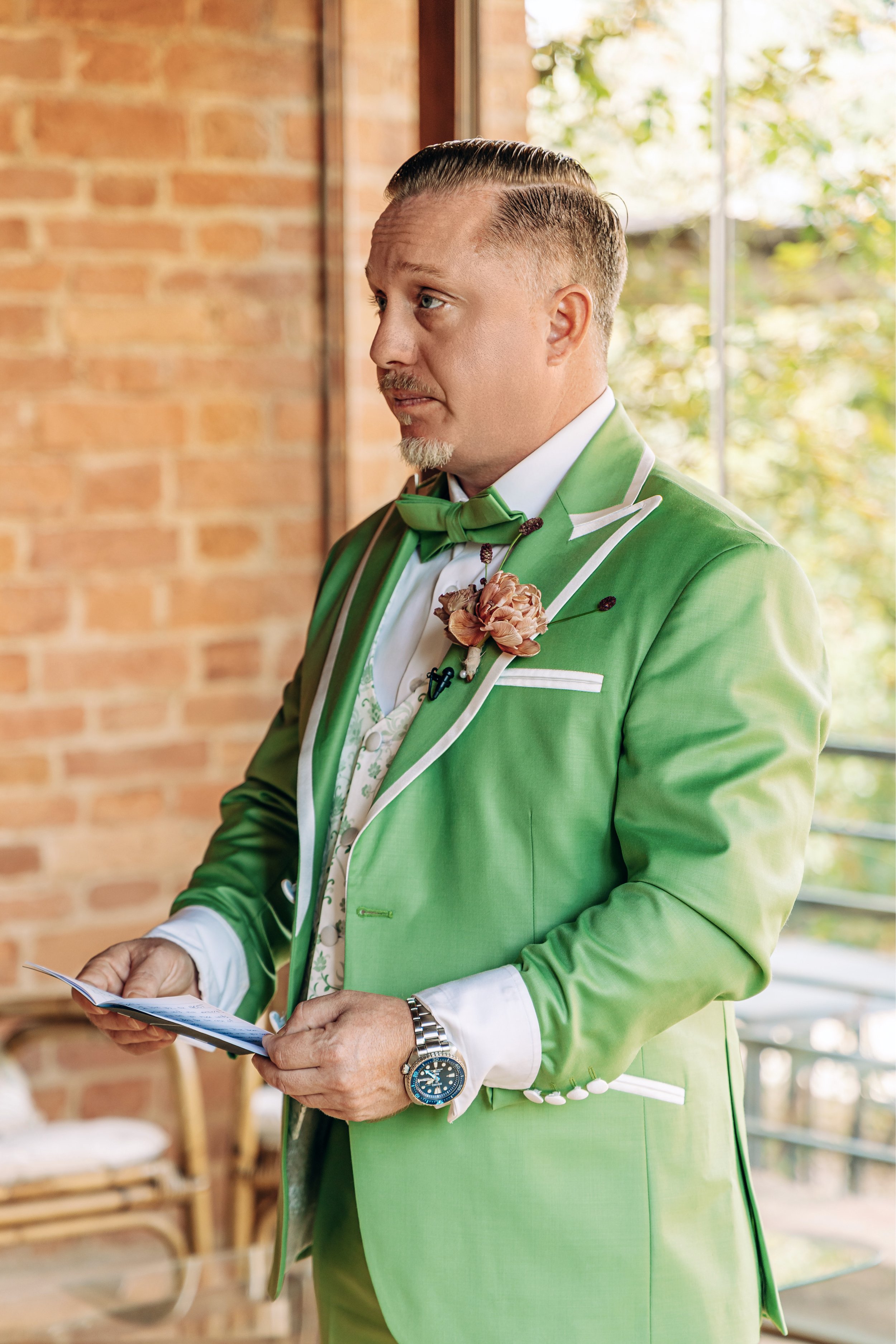 A man in a bright green tuxedo with a matching bow tie and boutonniere is holding a piece of paper and standing indoors near a window with a brick wall behind him. He is wearing a wristwatch and appears to be reading or reviewing something.