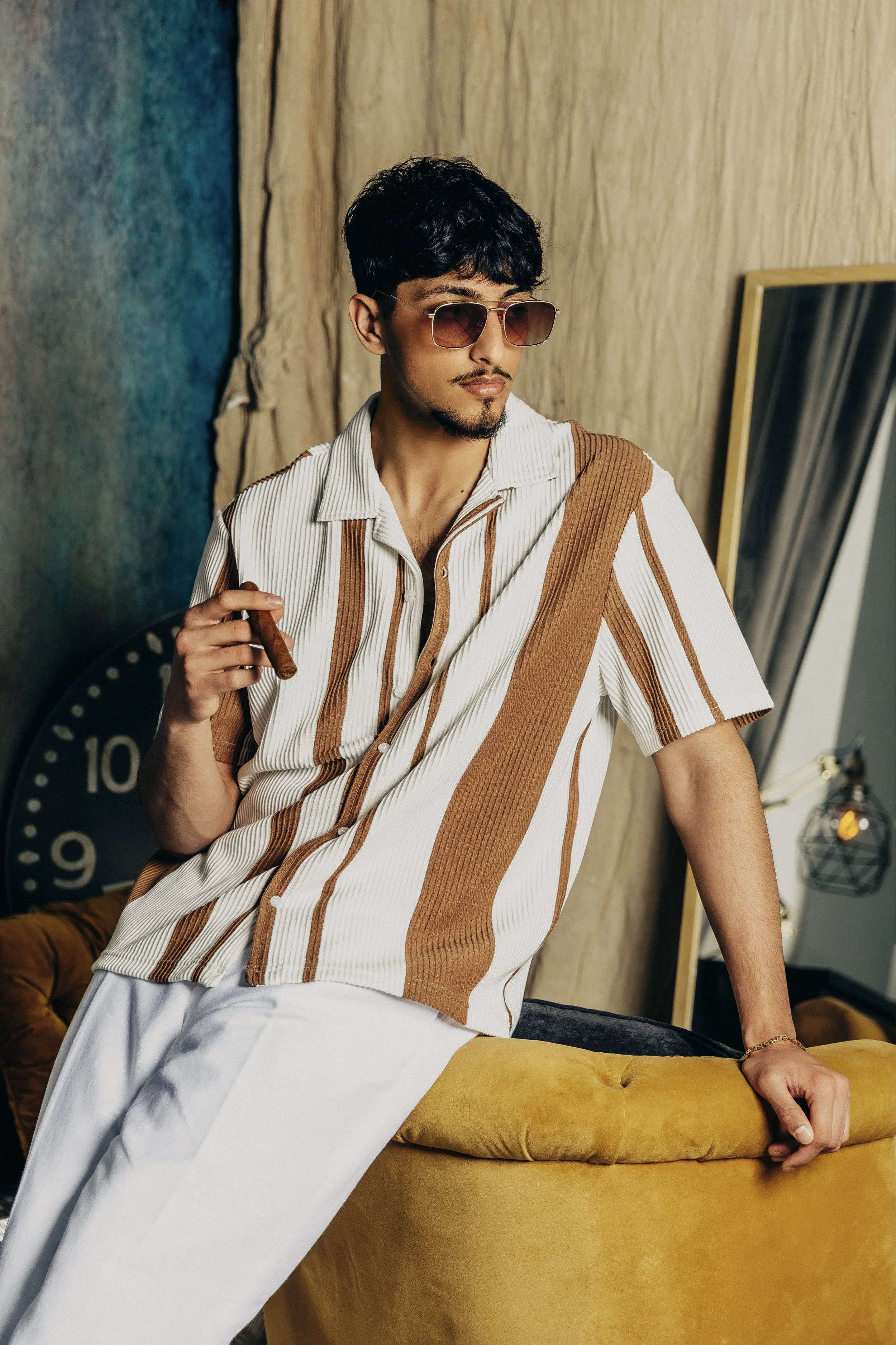 A stylish man with dark hair and sunglasses posing indoors with a cigar in his hand, leaning on a yellow velvet couch.