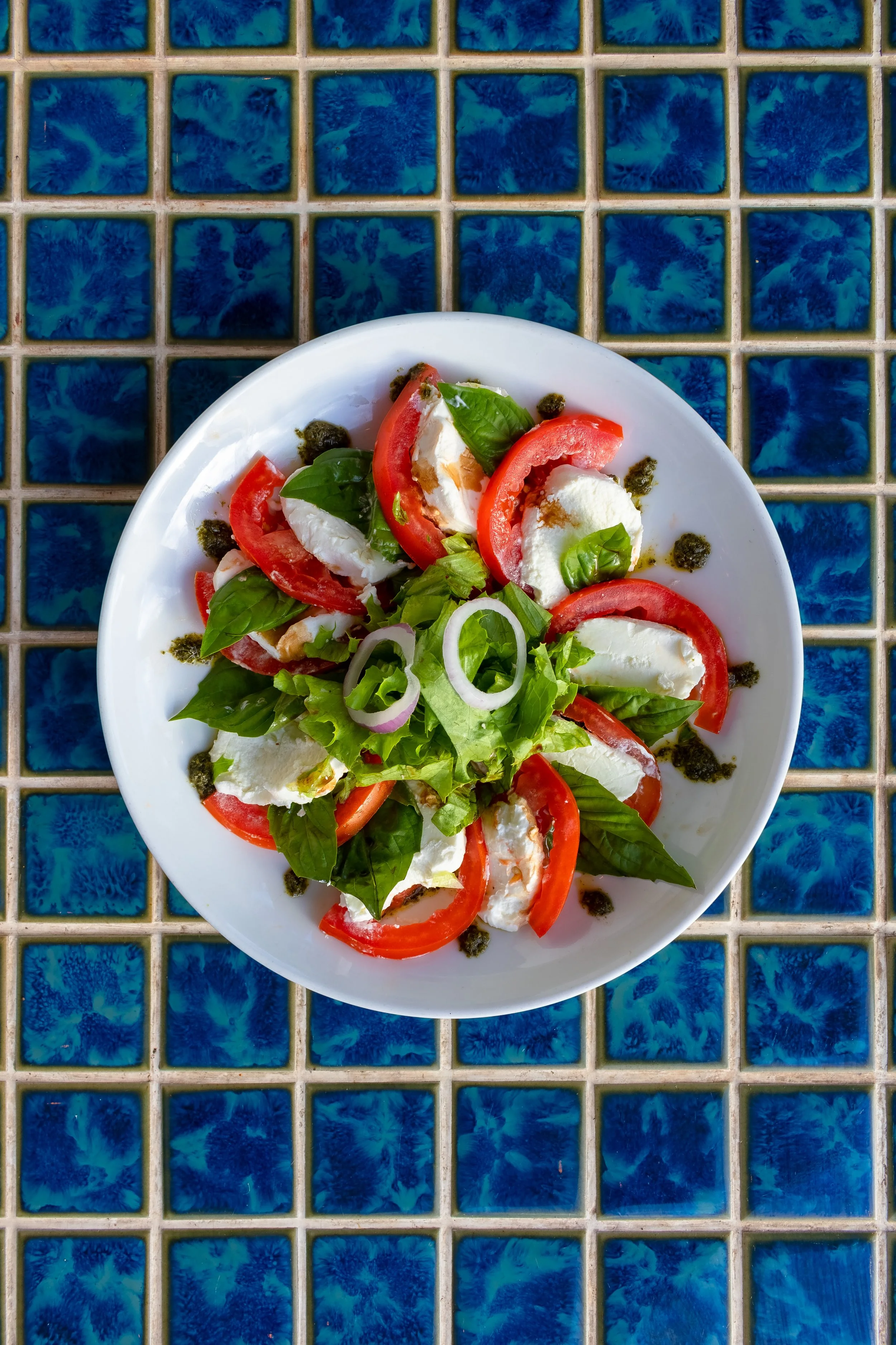 Caprese salad on white plate with tomato slices, fresh basil leaves, mozzarella cheese, and a drizzle of balsamic vinegar, placed on a blue tiled surface.