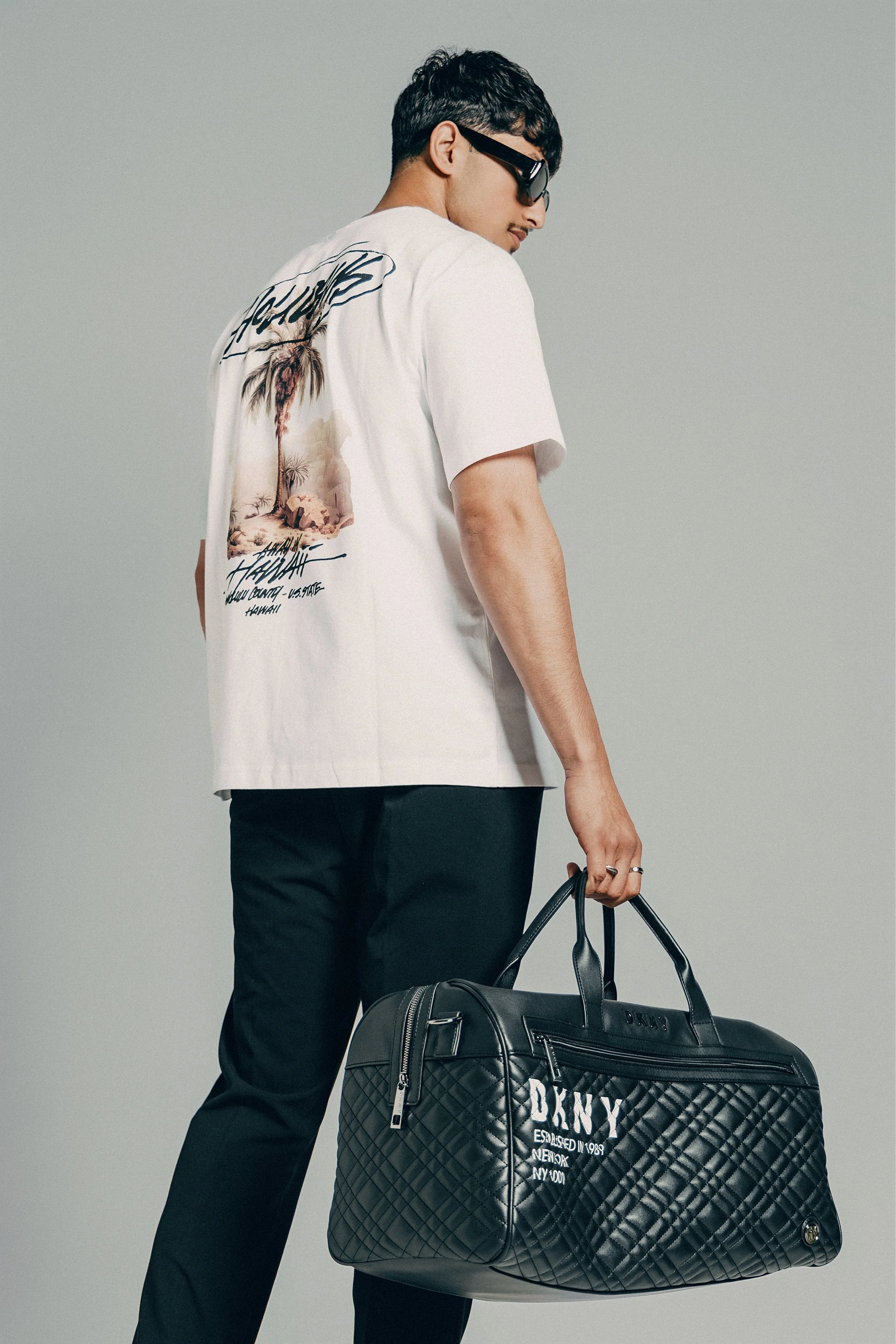 Man wearing sunglasses, white graphic t-shirt, and black pants, holding a large black quilted duffel bag against a plain gray background.